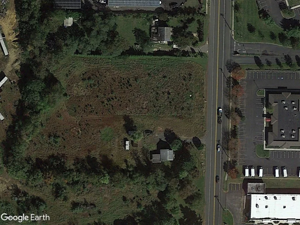Aerial view of a green irregularly shaped plot of land with surrounding trees, adjacent to a parking lot and buildings, and a road with cars driving on it.
