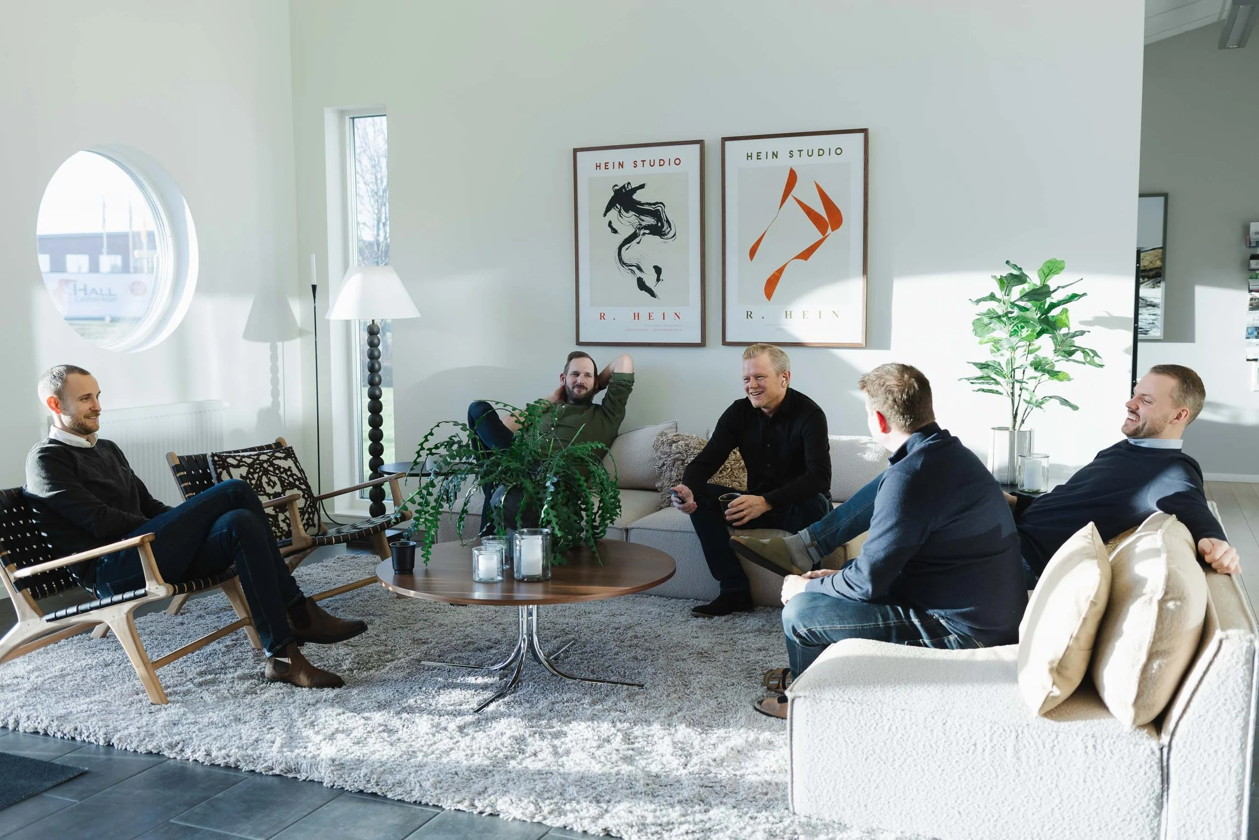 En grupp män som sitter och pratar i ett modernt vardagsrum med stora fönster, växt och konst på väggen.