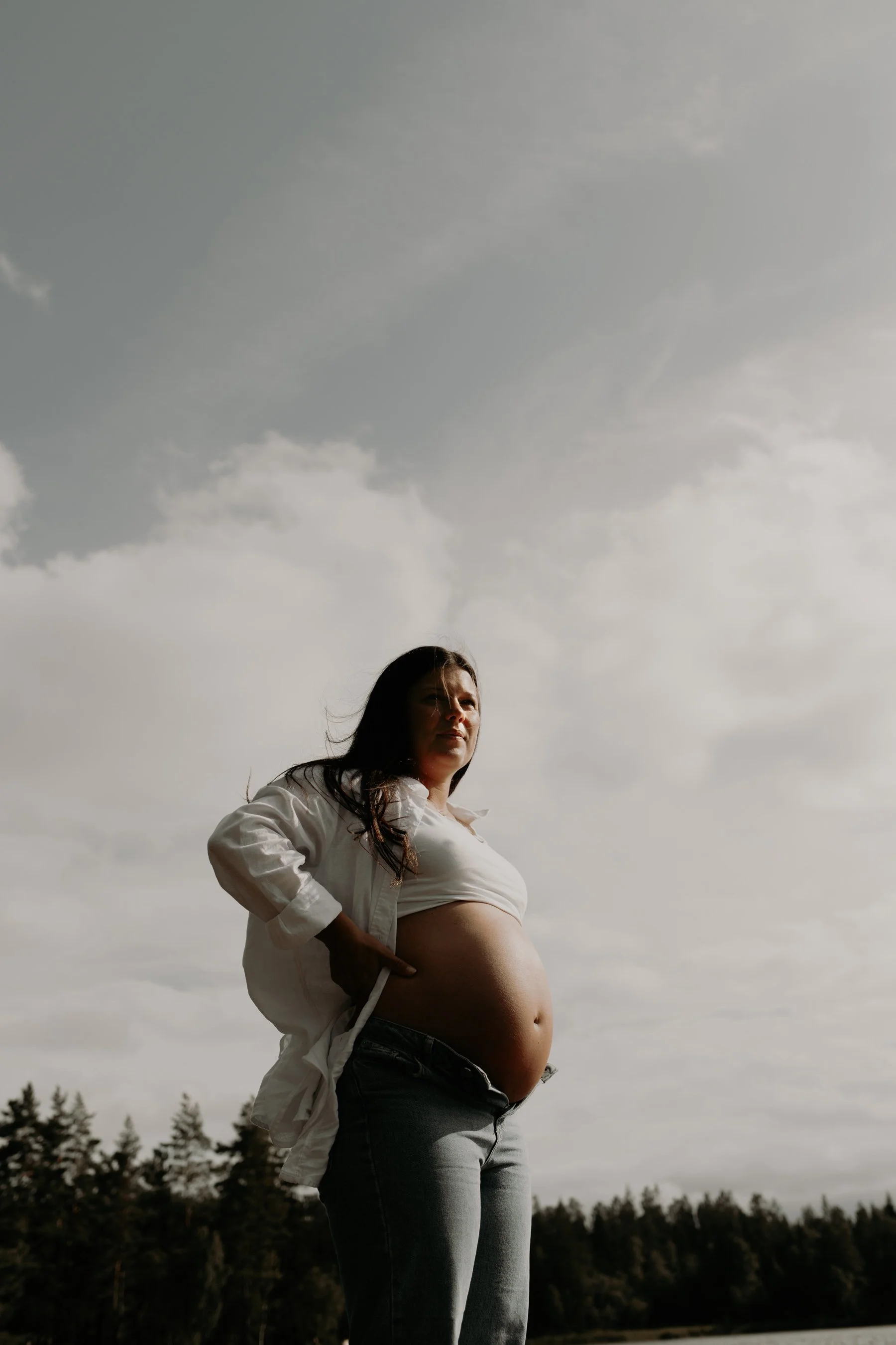 Gravid kvinna står utomhus med landskap av träd och himmel i bakgrunden.