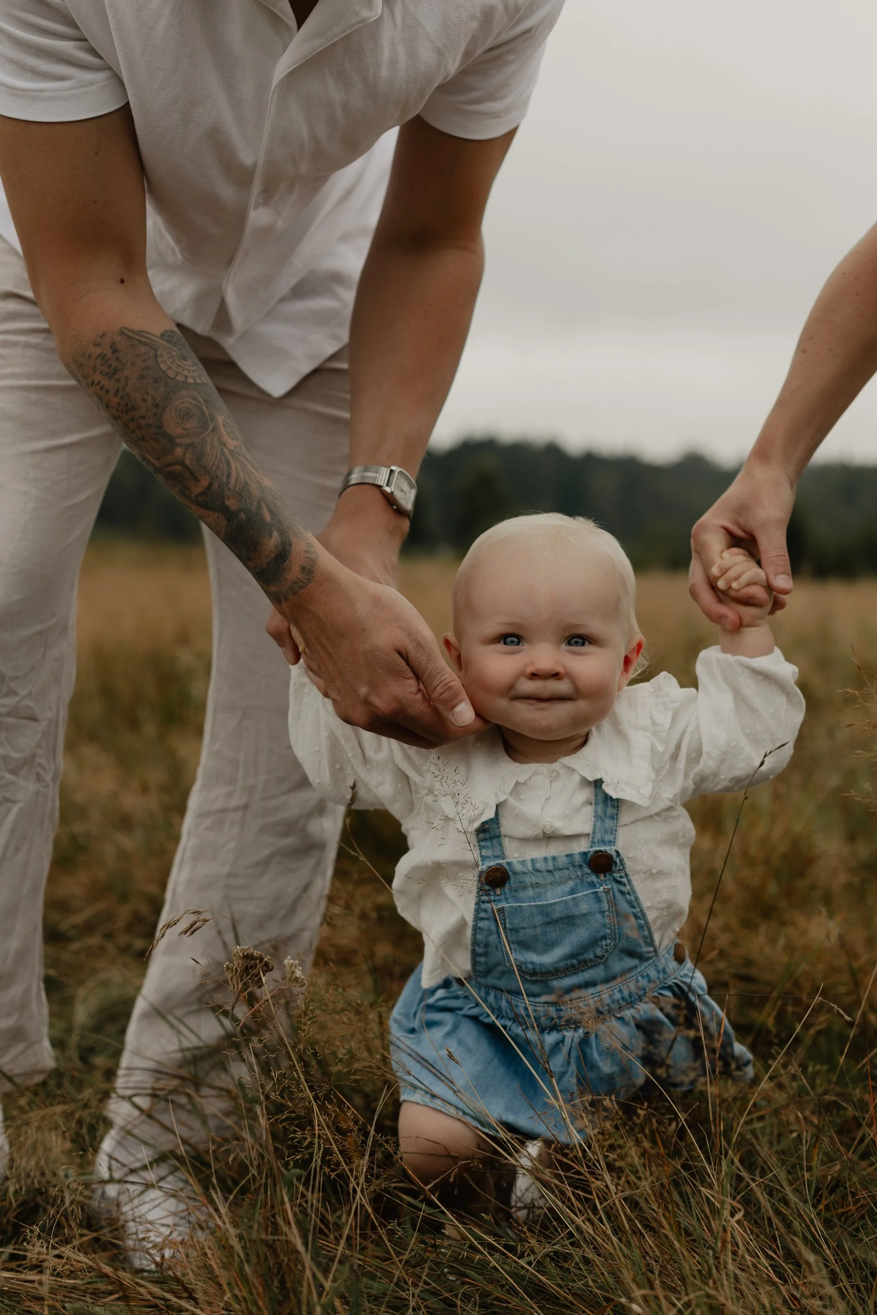 En vuxen person håller handen till ett barn som står i en åker. Barnet tittar mot kameran och ler. Personen är klädd i vit tröja och ljusa byxor, och har tatuering på armen.