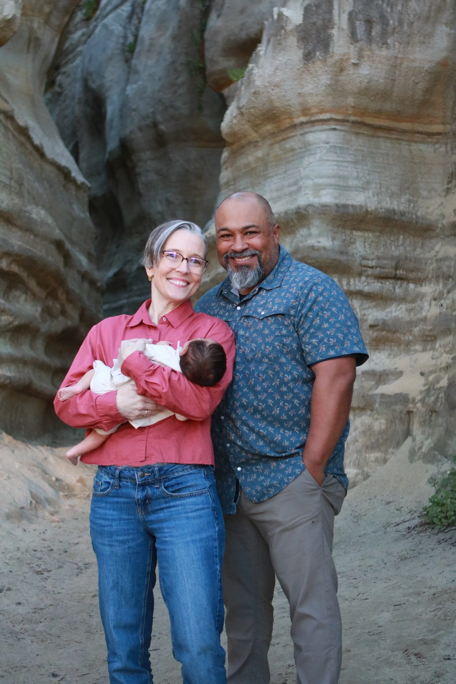 A smiling couple, a woman with glasses and short gray hair holding a newborn baby, standing outdoors with a rocky canyon background. The man, with a beard, wearing a blue patterned shirt, stands next to her.