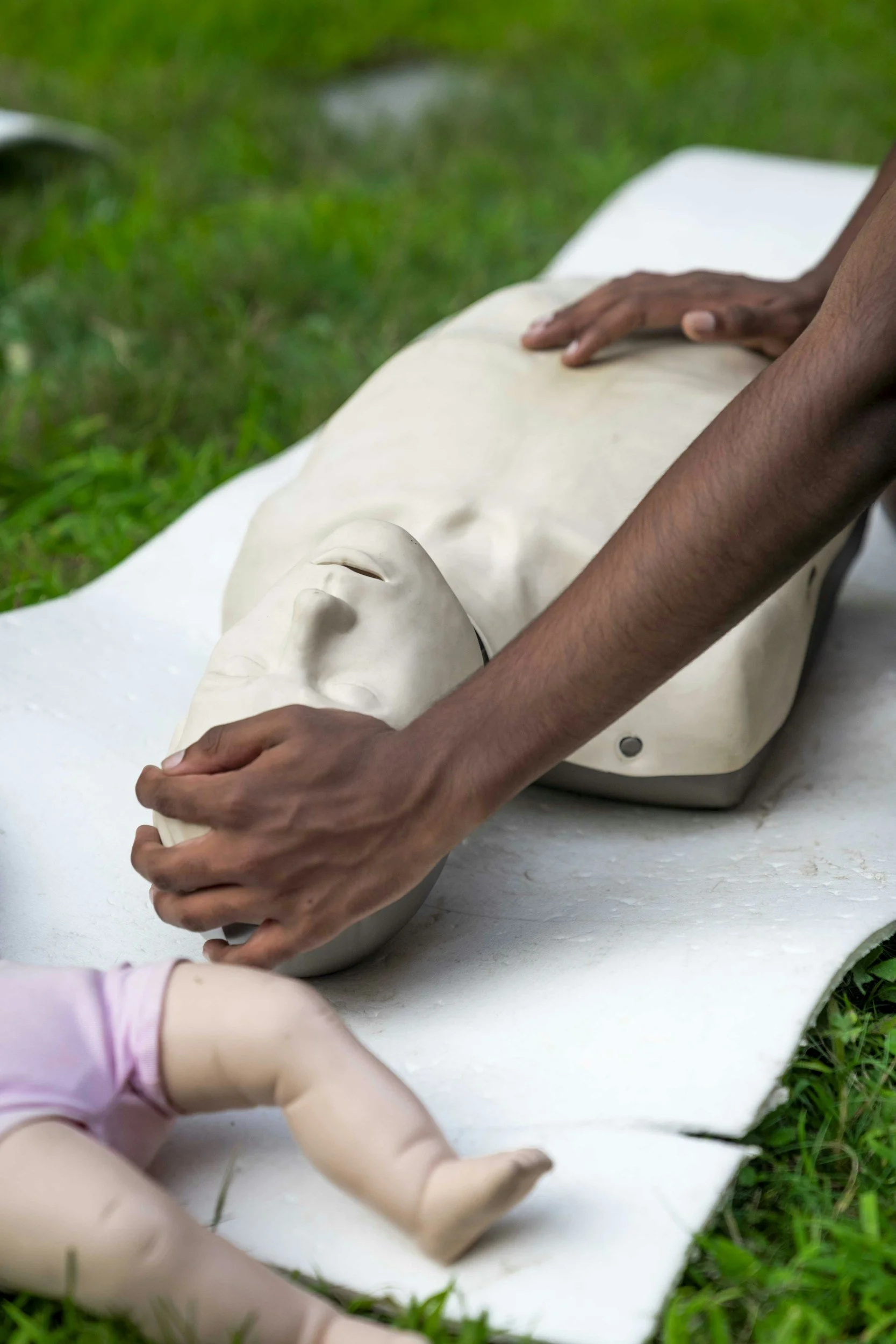 Person practicing CPR on a mannequin outdoors on a white surface, surrounded by green grass.