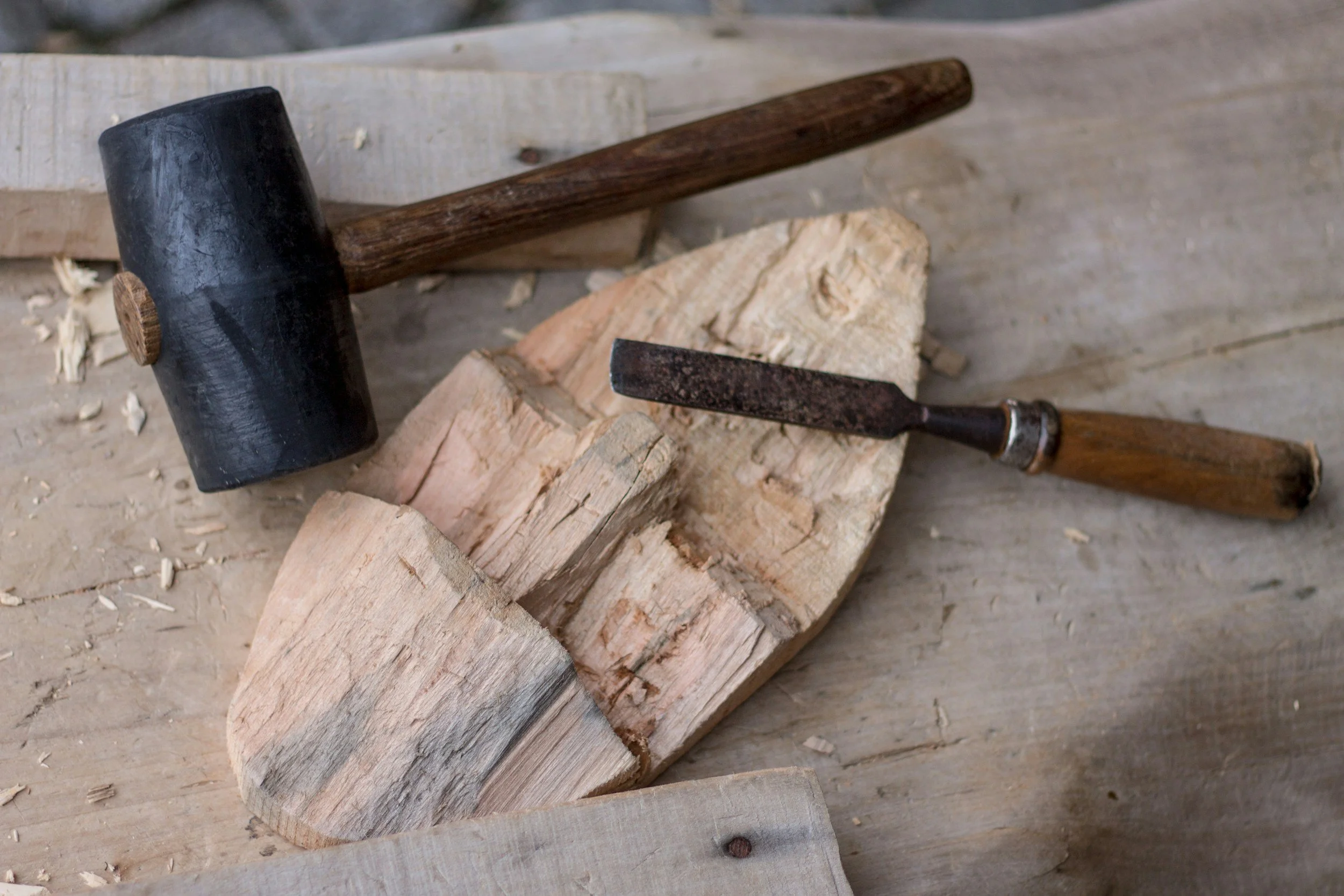 A hammer and chisel resting on chopped wood, with wood shavings scattered around on a wooden surface.