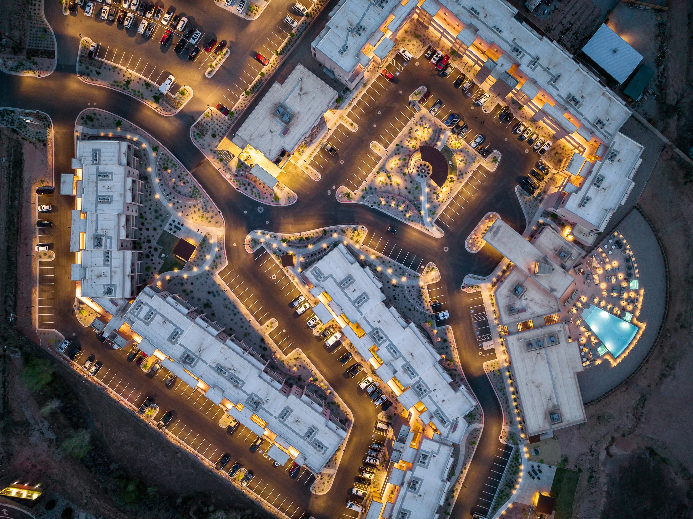 An aerial view of a multi-story apartment complex at dusk, showing parking lots, cars, and well-lit walkways.
