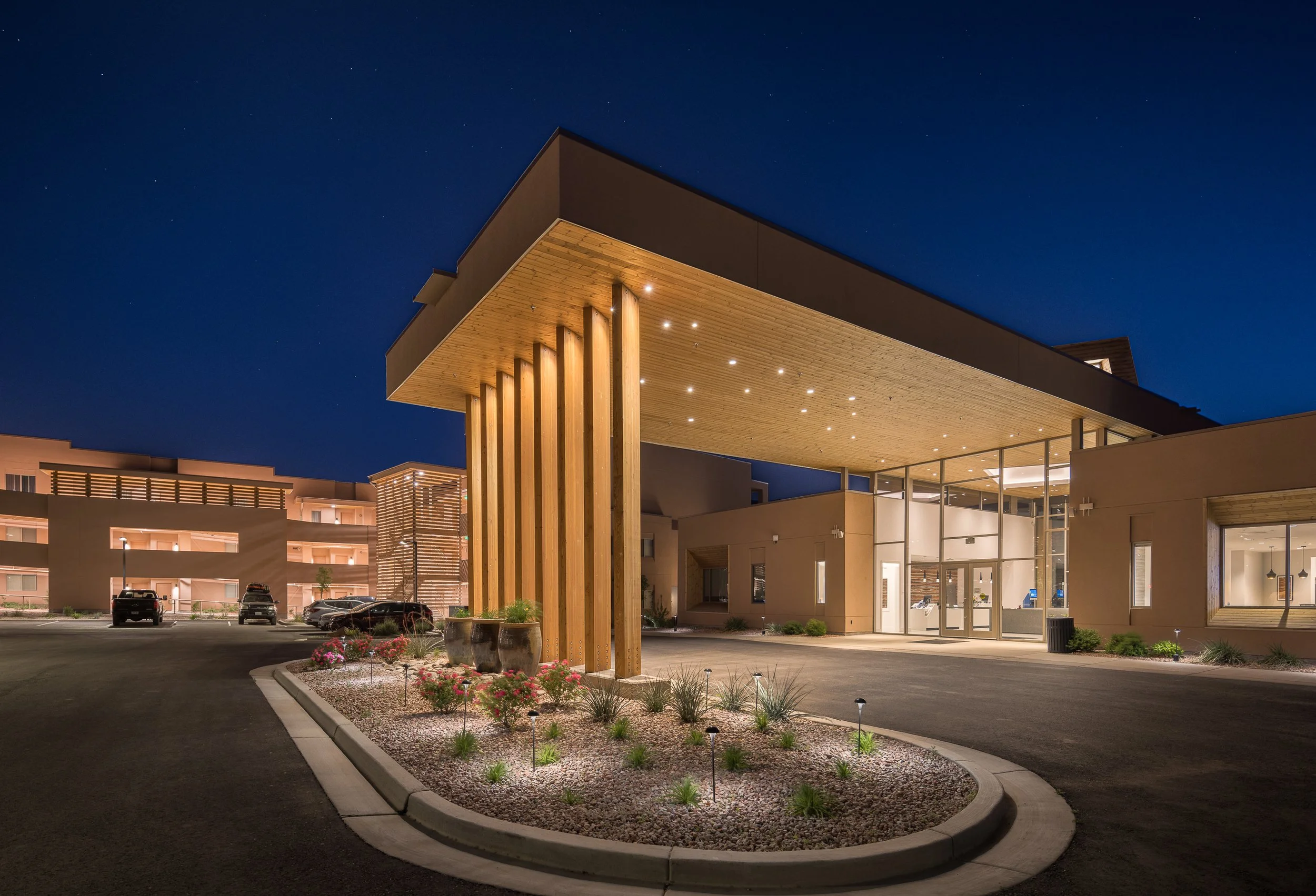 Night view of a modern building with a large underhang supported by wooden beams, illuminated outdoor lights, a landscaped area with small plants and flowers, parked cars, and a clear night sky with stars.