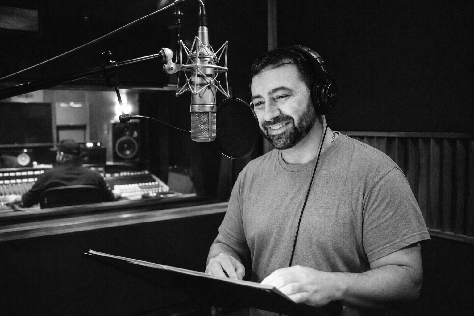A man in a recording studio wearing headphones, smiling, standing in front of a microphone with a pop filter, with a sound engineer working at a mixing console in the background.
