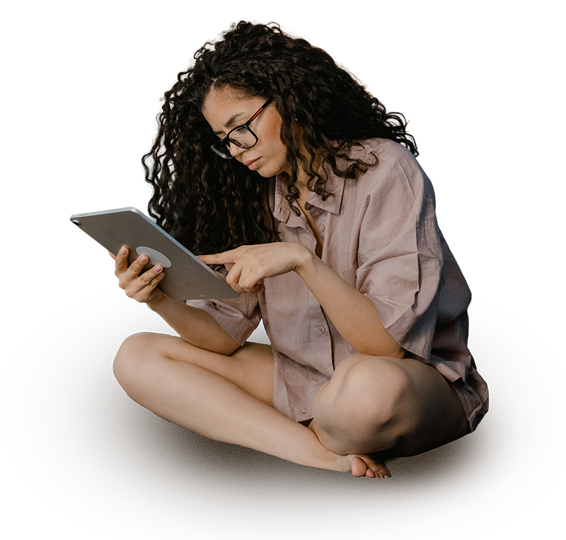 Young woman with curly hair and glasses sitting cross-legged on the floor, reading a tablet