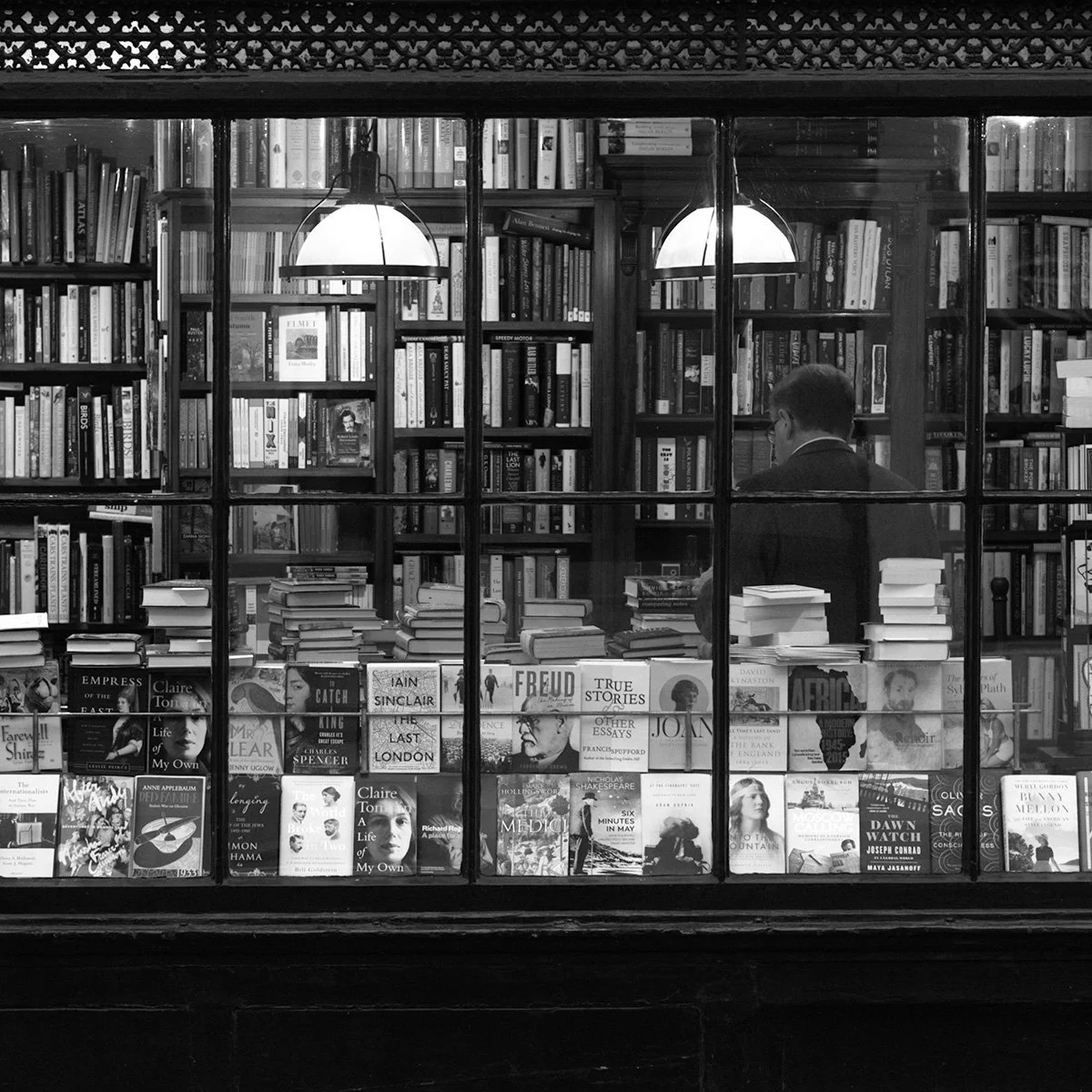 View of a bookstore through a glass window with bookshelves, a person reading, and a table of books in the foreground.