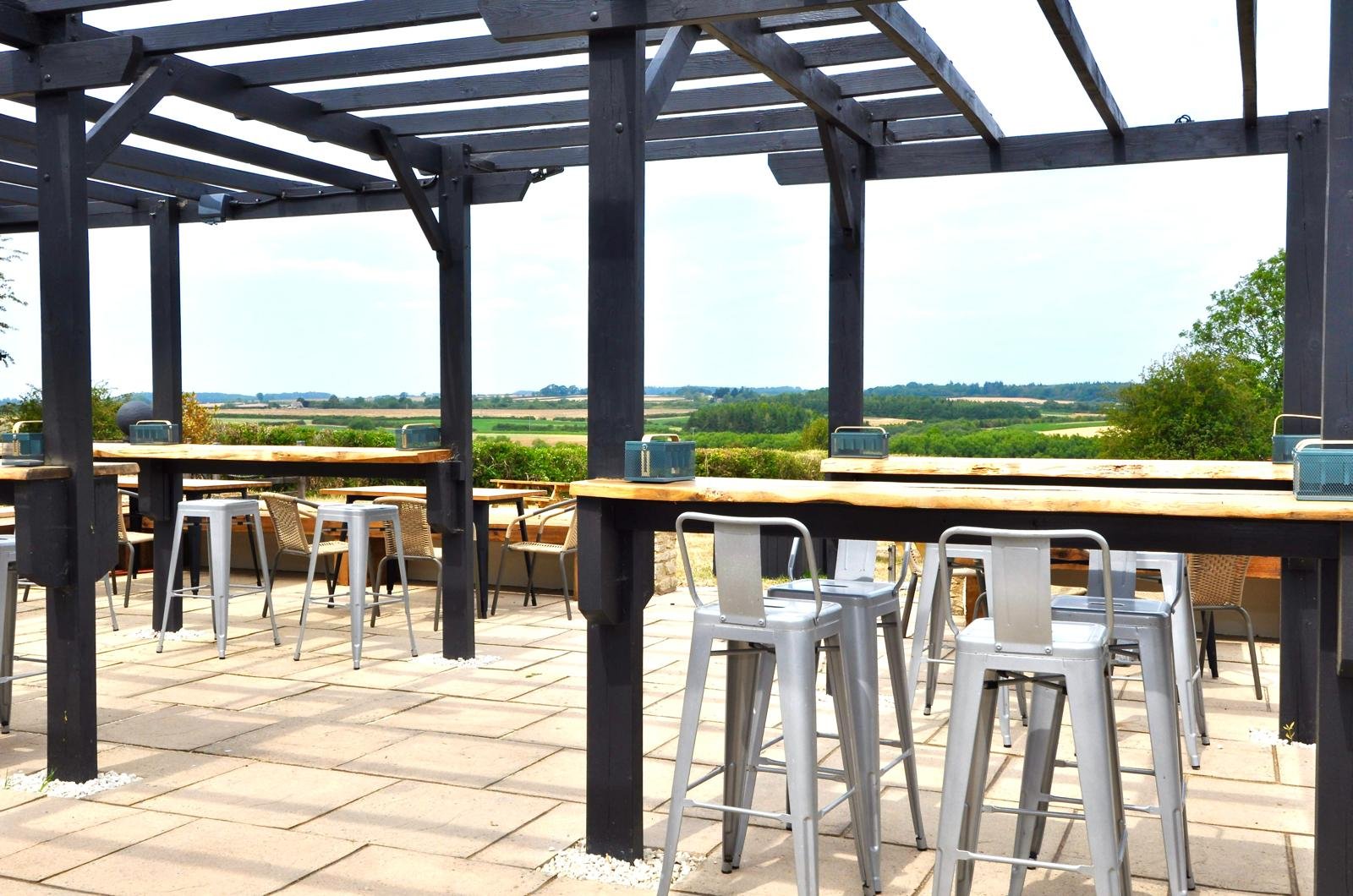 Outdoor patio with high tables and metal bar stools, and a wooden pergola, overlooking a green landscape with fields and trees.