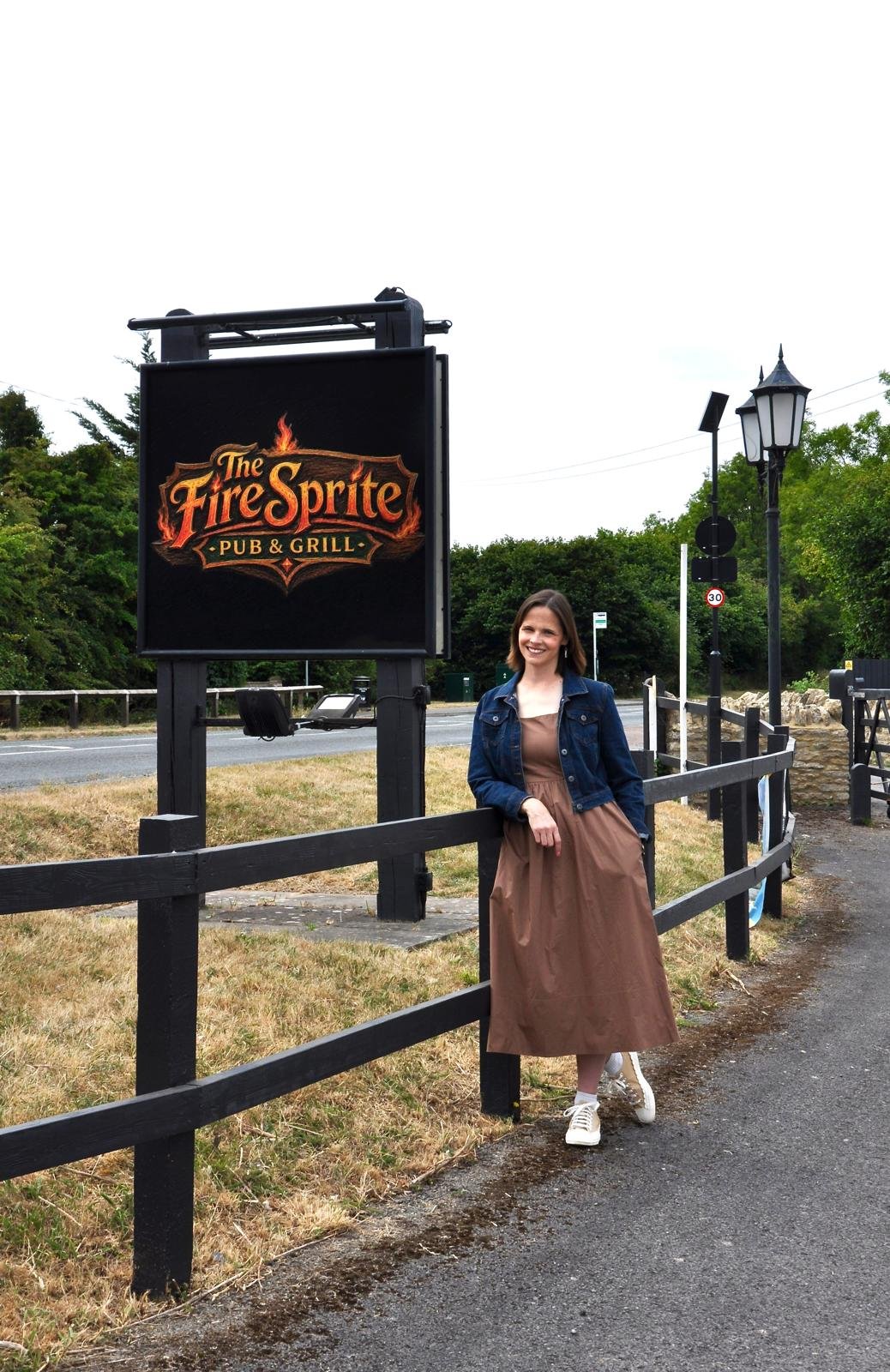 A woman in a brown dress and denim jacket standing next to a black wooden fence on a sidewalk, smiling, with a sign for The Fire Sprite Pub & Grill and green trees in the background.