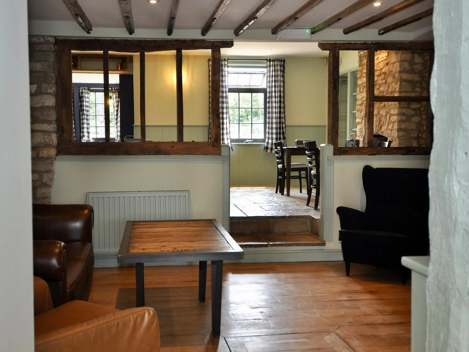 Interior of a rustic restaurant or cafe with a wooden and brick design, featuring a small seating area, tables, and chairs, and a split-level floor with a window and checkered curtains.