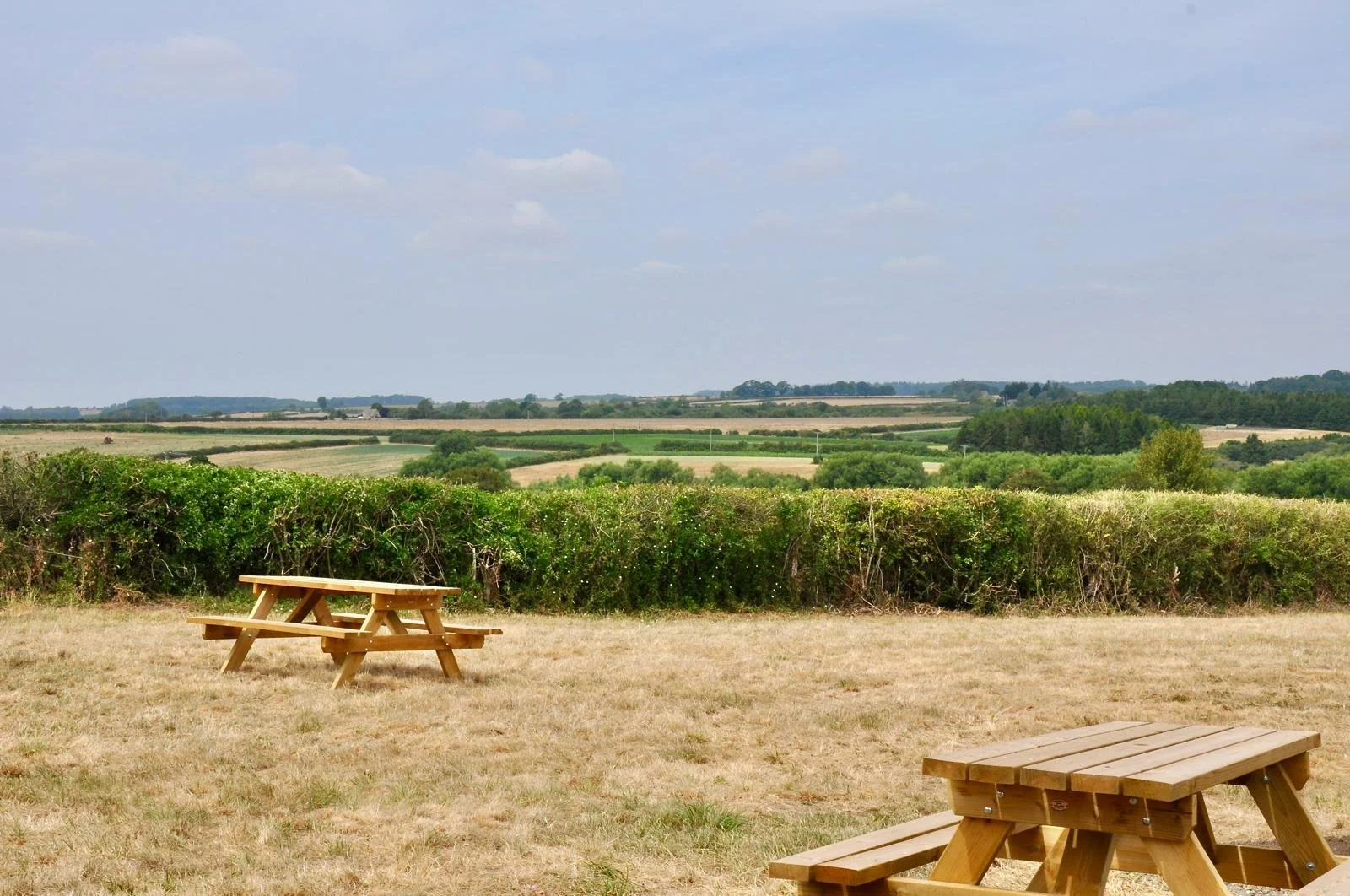 Two wooden picnic tables on a grassy field overlooking a countryside landscape with rolling fields and a hedge in the background under a partly cloudy sky.