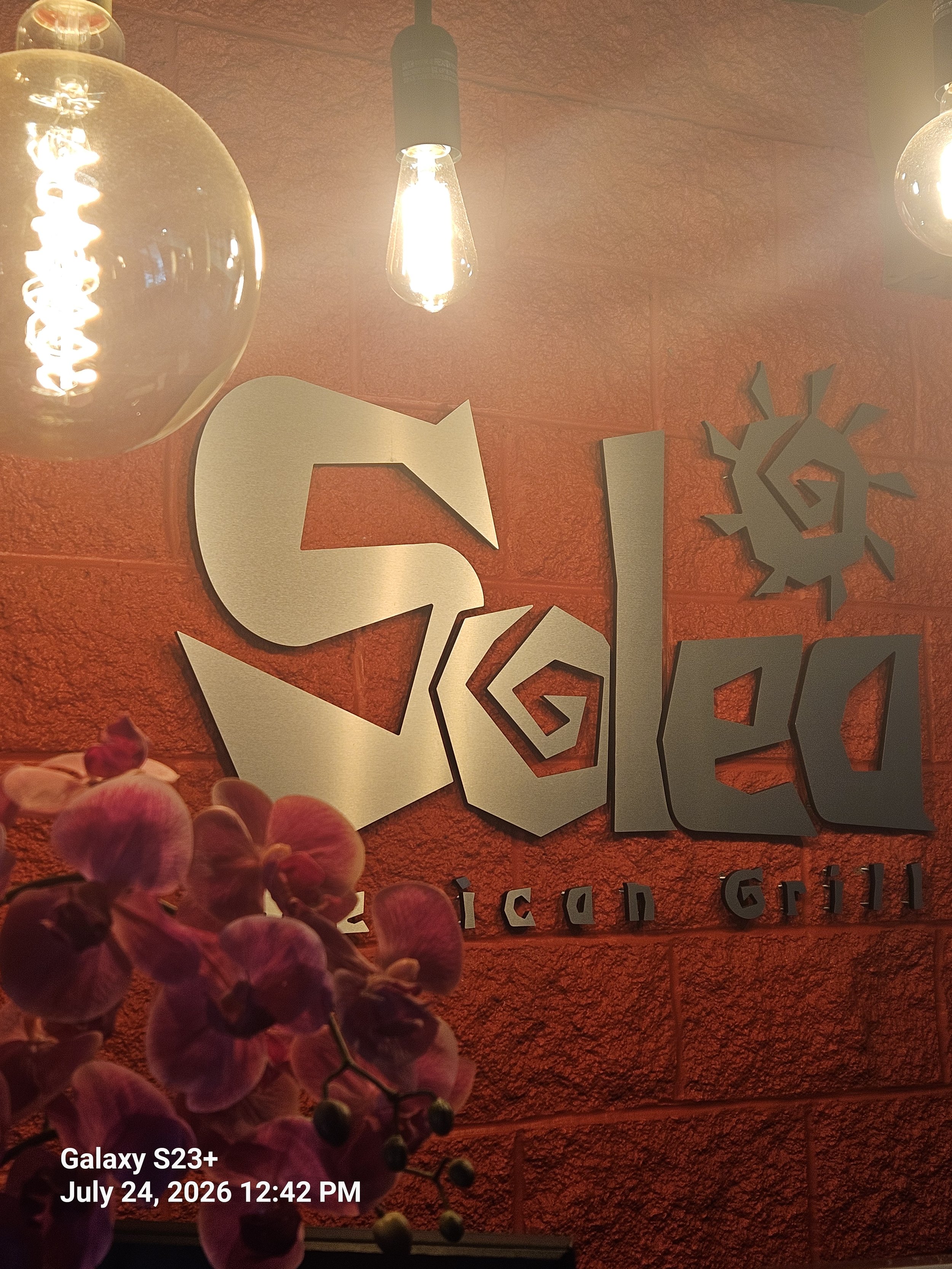 Close-up of decorative wall with metallic letters spelling 'SIX' and geometric design, orange textured wall, table lamp with exposed filament bulb, pink orchids in foreground, timestamp in lower left corner indicating photo was taken on July 24, 2026, at 12:42 PM.