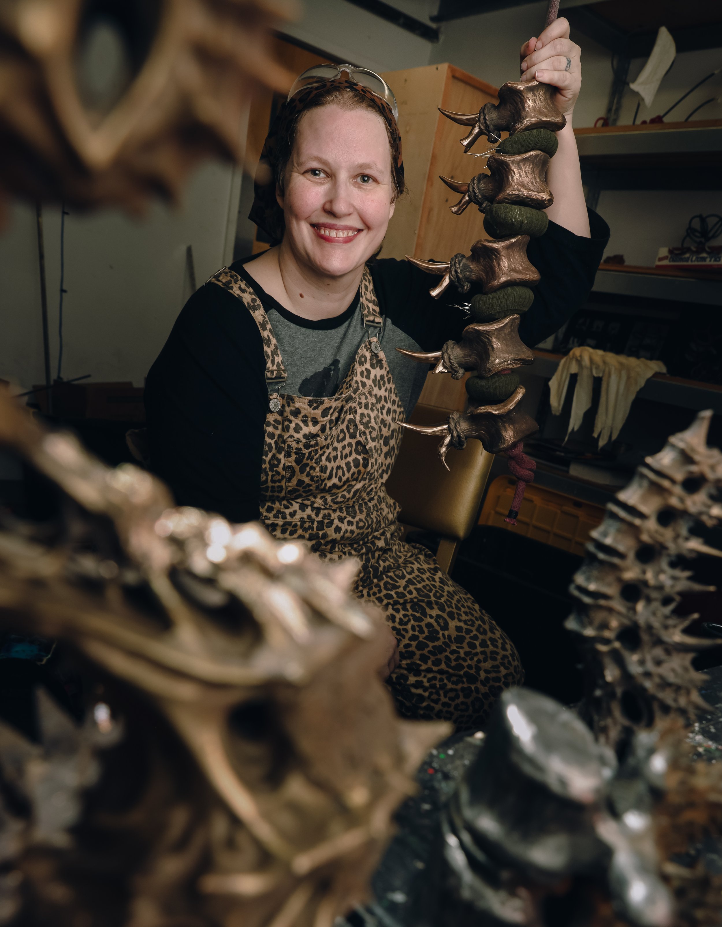 A woman smiling and holding a large bronze sculpture of a spine suspended.  With other bronze sculptures surrounding her in an art studio