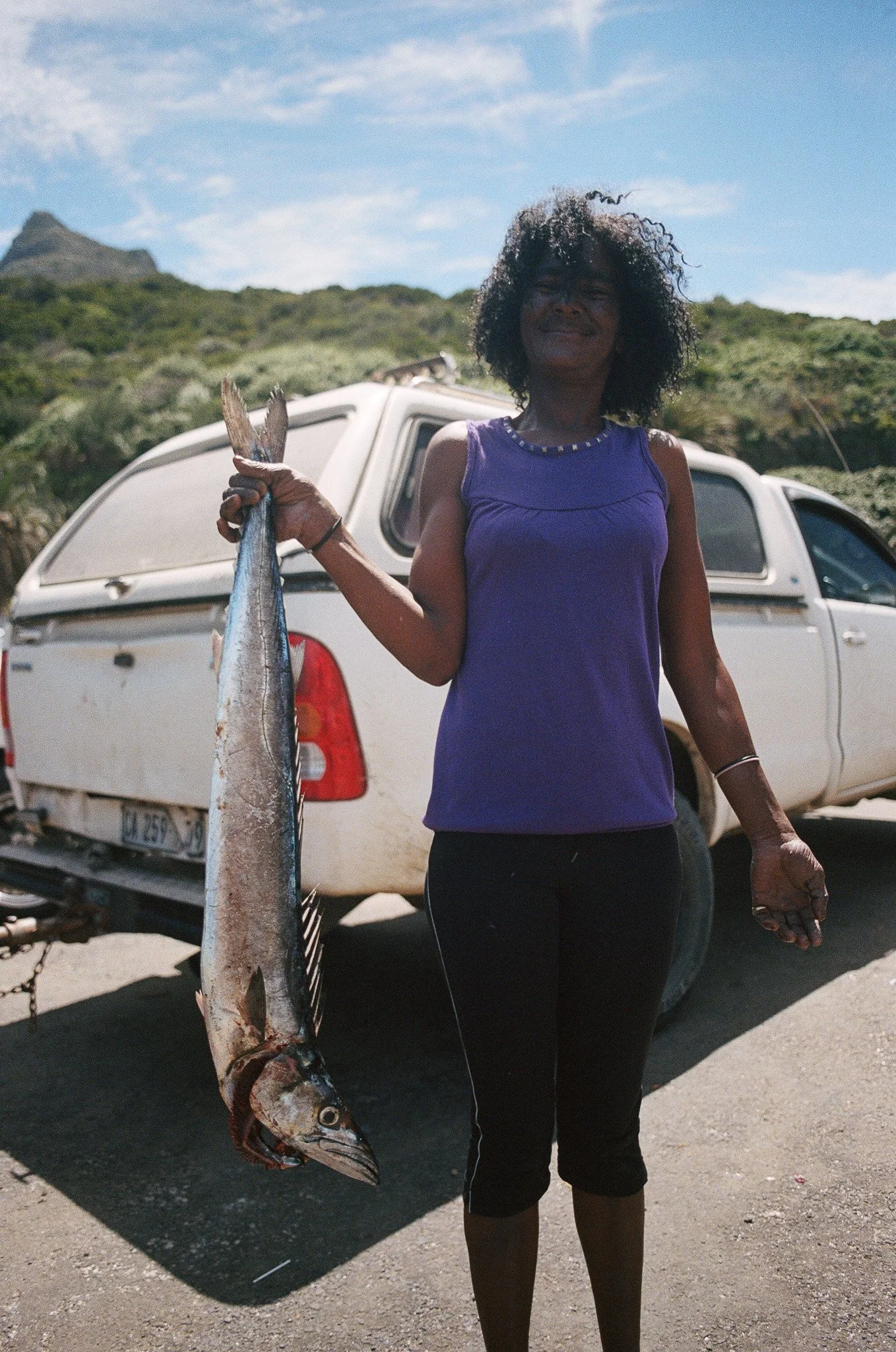 Woman in a purple sleeveless top holding a large fish in front of a white pickup truck on a sunny day, with green hills and a partly cloudy sky in the background.