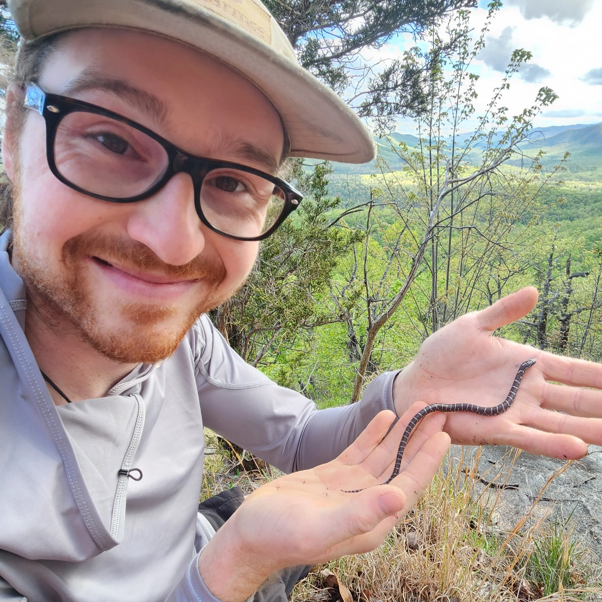 A person with glasses and a hat holding a snake in an outdoor natural setting with trees and mountains in the background.