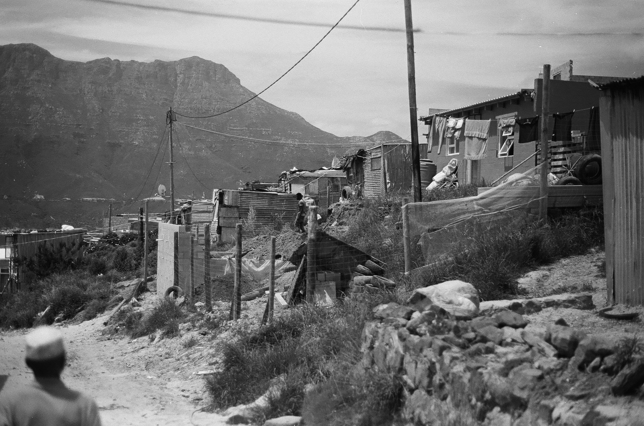 A black and white photo of a rural area with makeshift houses made of scrap metal, laundry hanging on lines, utility poles with power lines, rocks, and a mountain in the background.