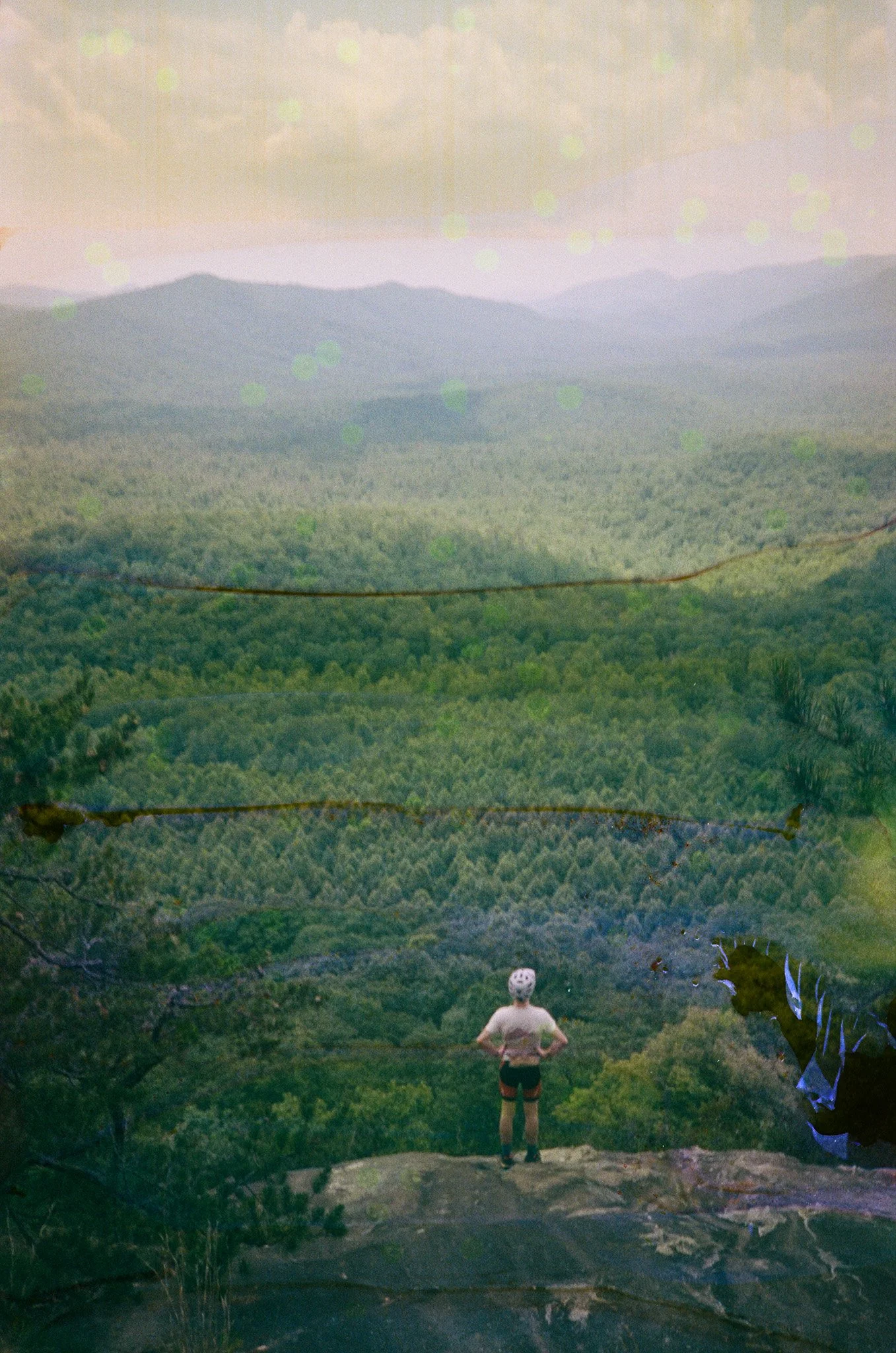 A person with hands on hips standing on a rocky ledge, overlooking a vast, green forested valley with distant mountain ranges under a cloudy sky.