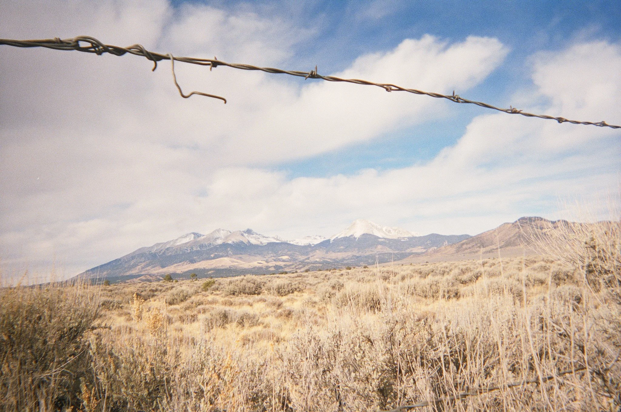 A barbed wire fence in a dry grass field with snow-capped mountains and a partly cloudy sky in the background.