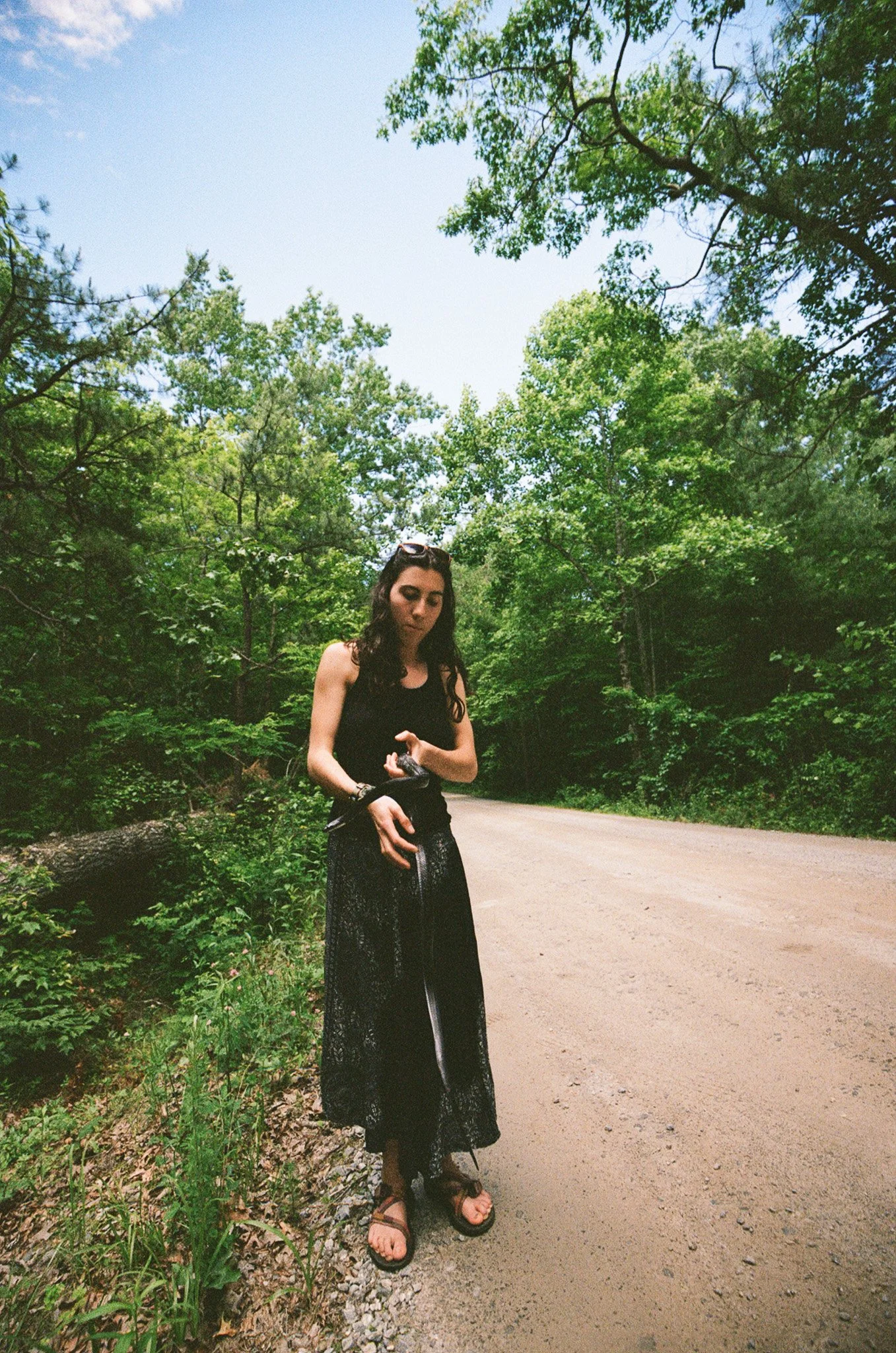 A woman with long dark hair standing on a dirt path in a lush green forest, wearing a black sleeveless top and a long black skirt, with sunglasses on her head, and holding a camera in her hand.
