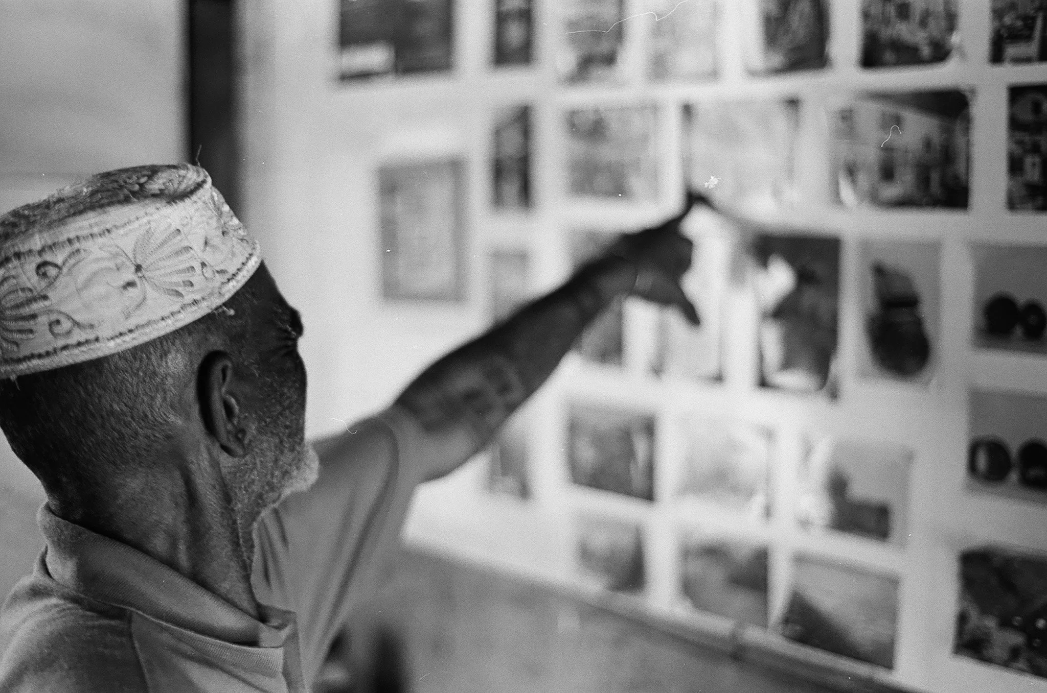 A person wearing a patterned cap is reaching out and pointing towards a wall filled with photographs or artwork.