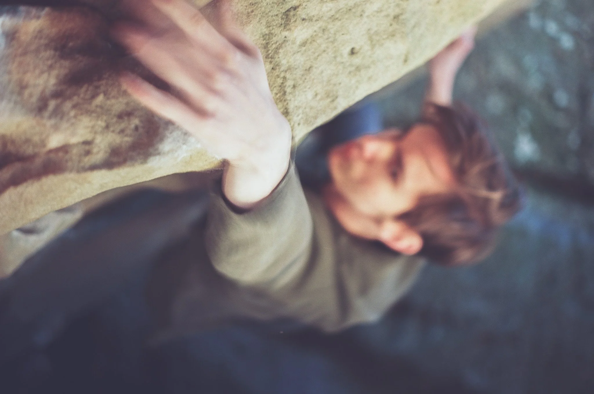 A person with short brown hair is lying down on a surface, looking up at the camera. The person's hand is resting on their chest, and they are wearing a dark top. The background includes a beige or tan surface, possibly a carpet or blanket, with blur