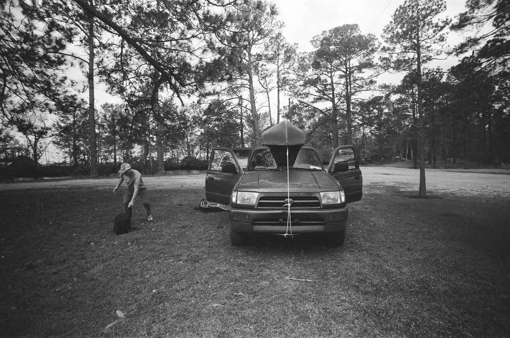 A black and white image of a car with the hood open in a forest clearing, with a man in a cap working near the car and tall trees in the background.
