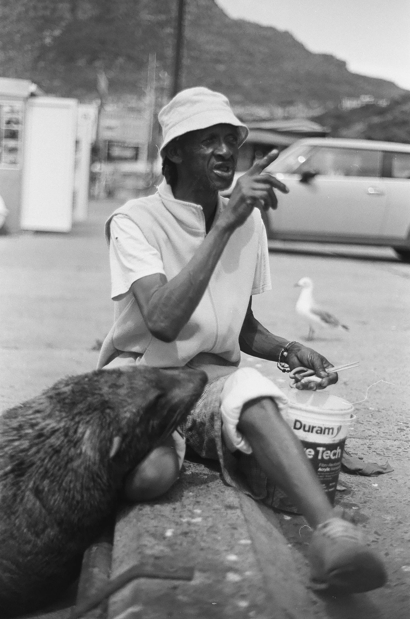 A black-and-white photo of a man sitting on the ground, wearing a bucket hat and a short-sleeved shirt, gesturing with his index finger, with a seagull nearby and mountains in the background.