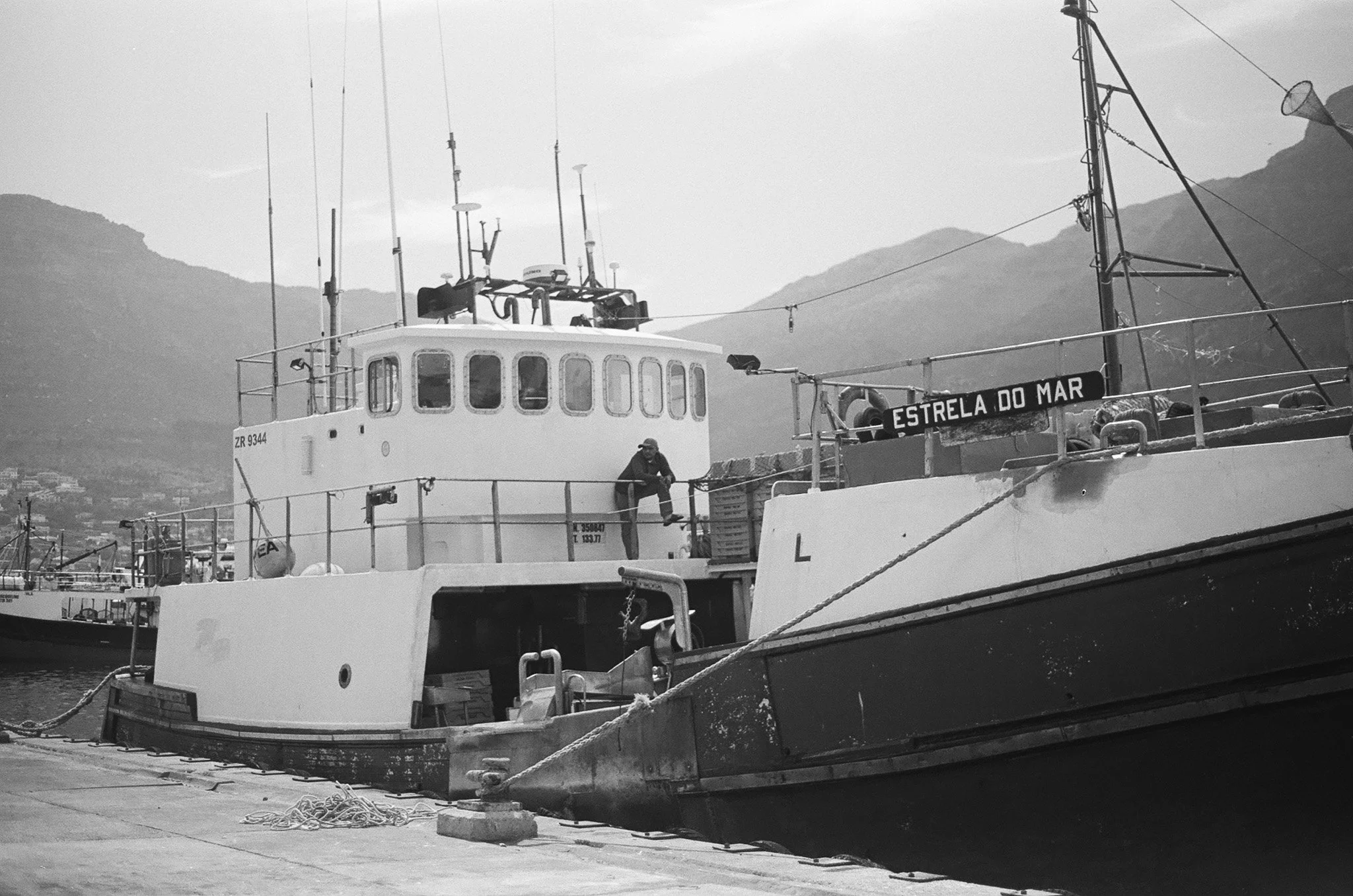 Black and white photo of boats docked at a harbor, with a mountain range in the background and a person sitting on the boat's deck near a sign that reads 'Estrela Do Mar'.