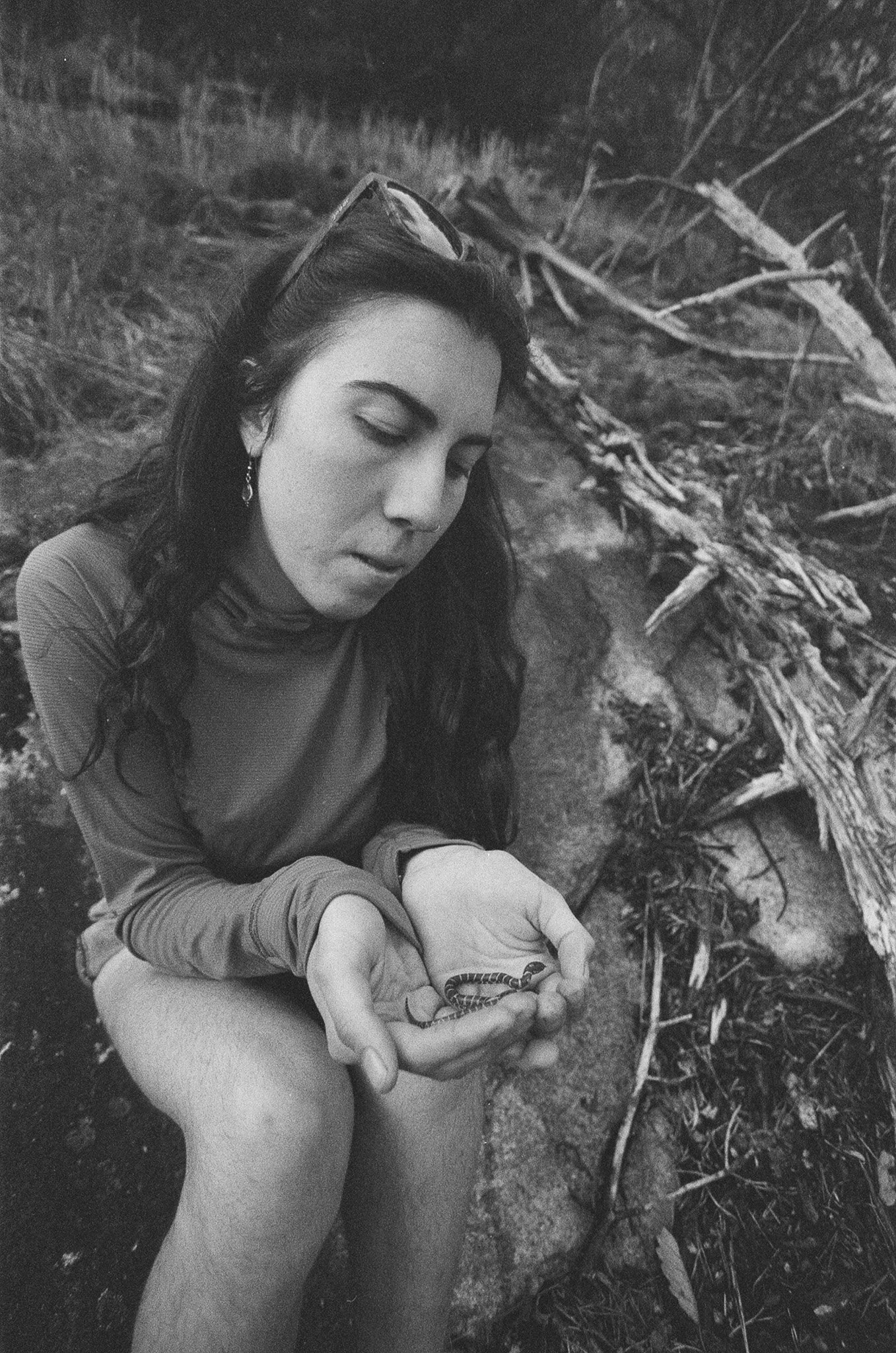 A woman with long dark hair, wearing a long-sleeved shirt, is sitting outdoors on the ground with her eyes closed, holding a small snake in her hands. The background features a fallen tree and natural outdoor scenery in black and white.