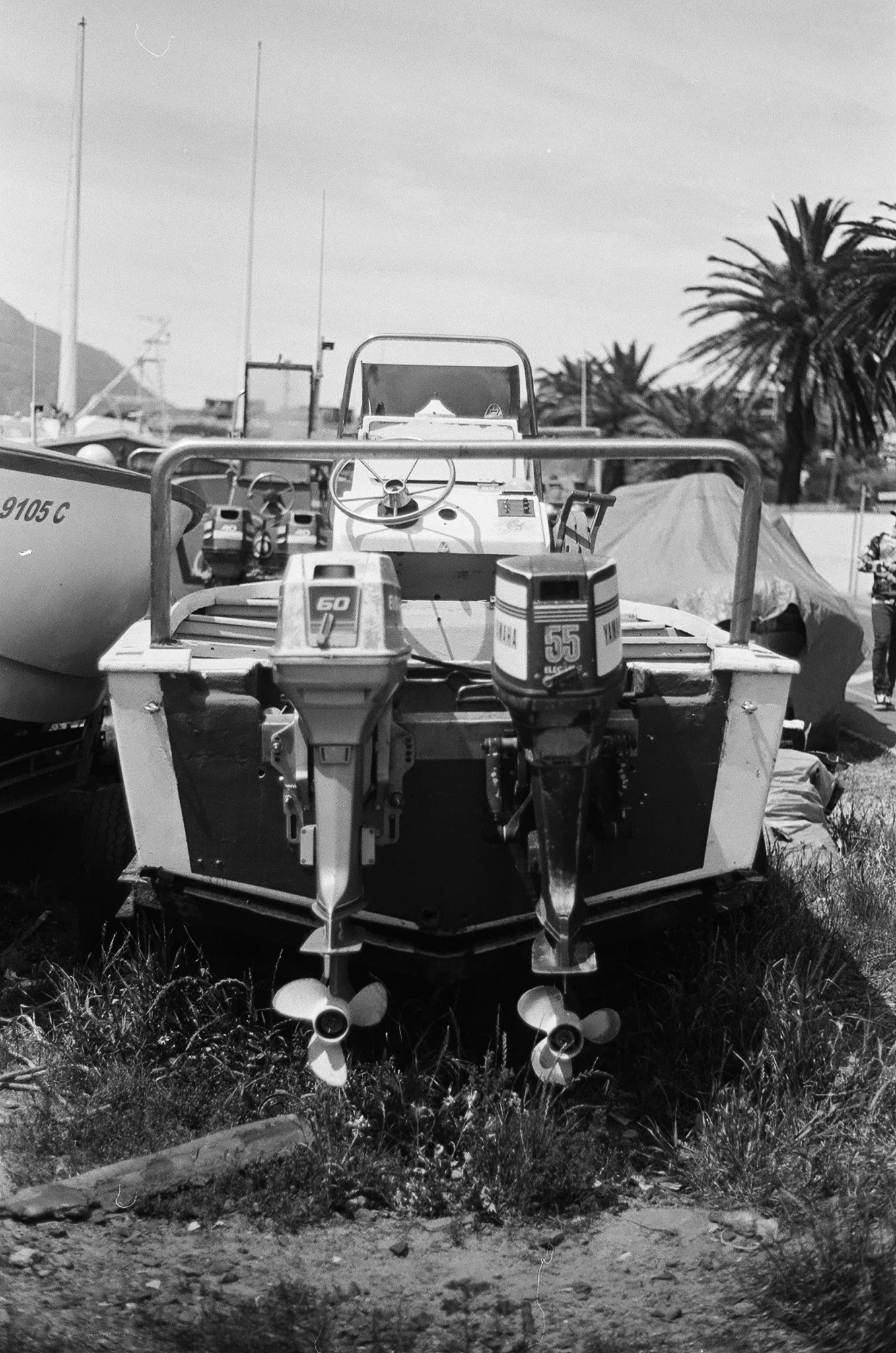 Black and white photo of a small boat with outboard motors, parked on grass in an outdoor area with trees and power lines in the background.