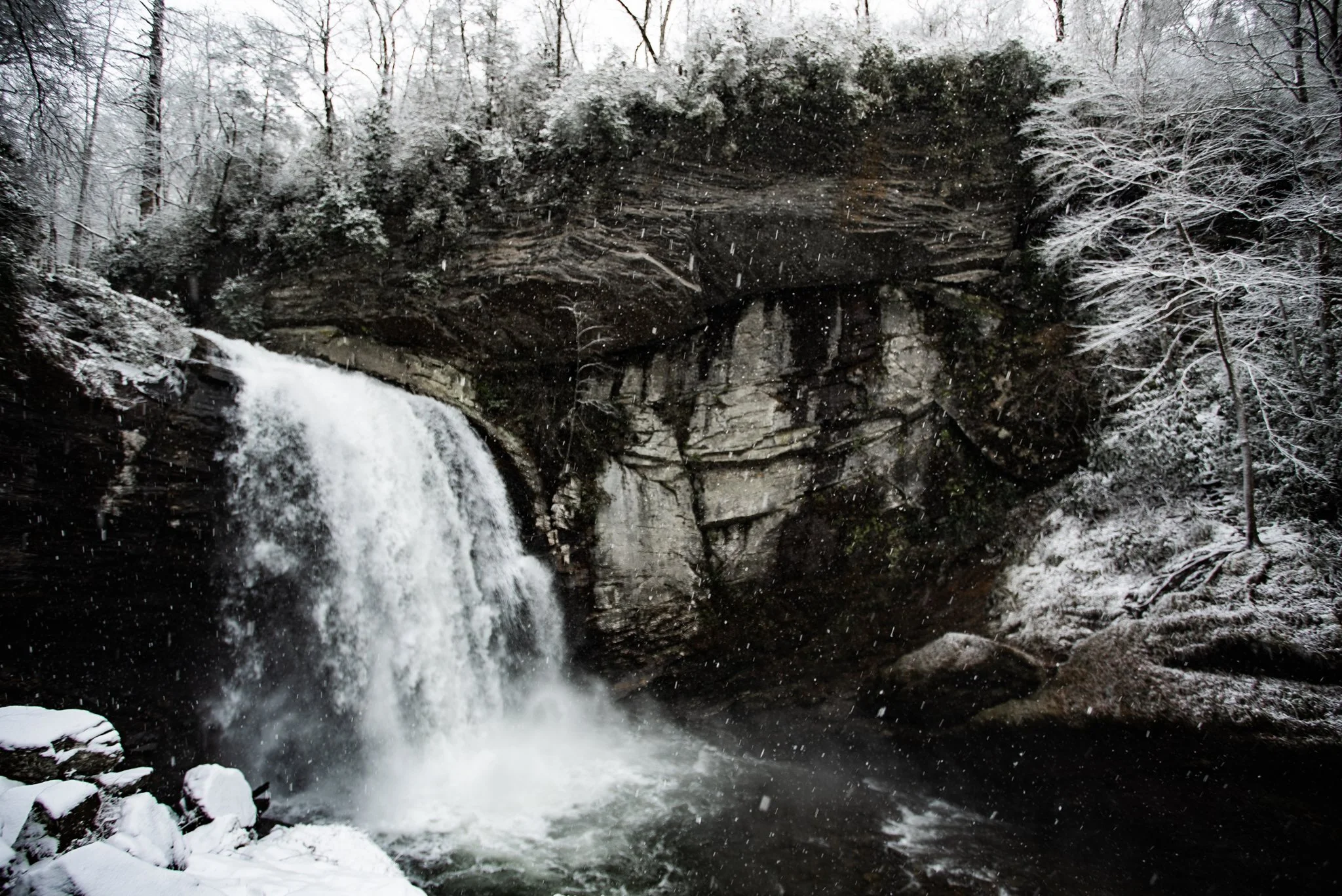 A small waterfall cascading down rocks into a river, surrounded by snow-covered trees and rocks, with snow falling in a winter landscape.