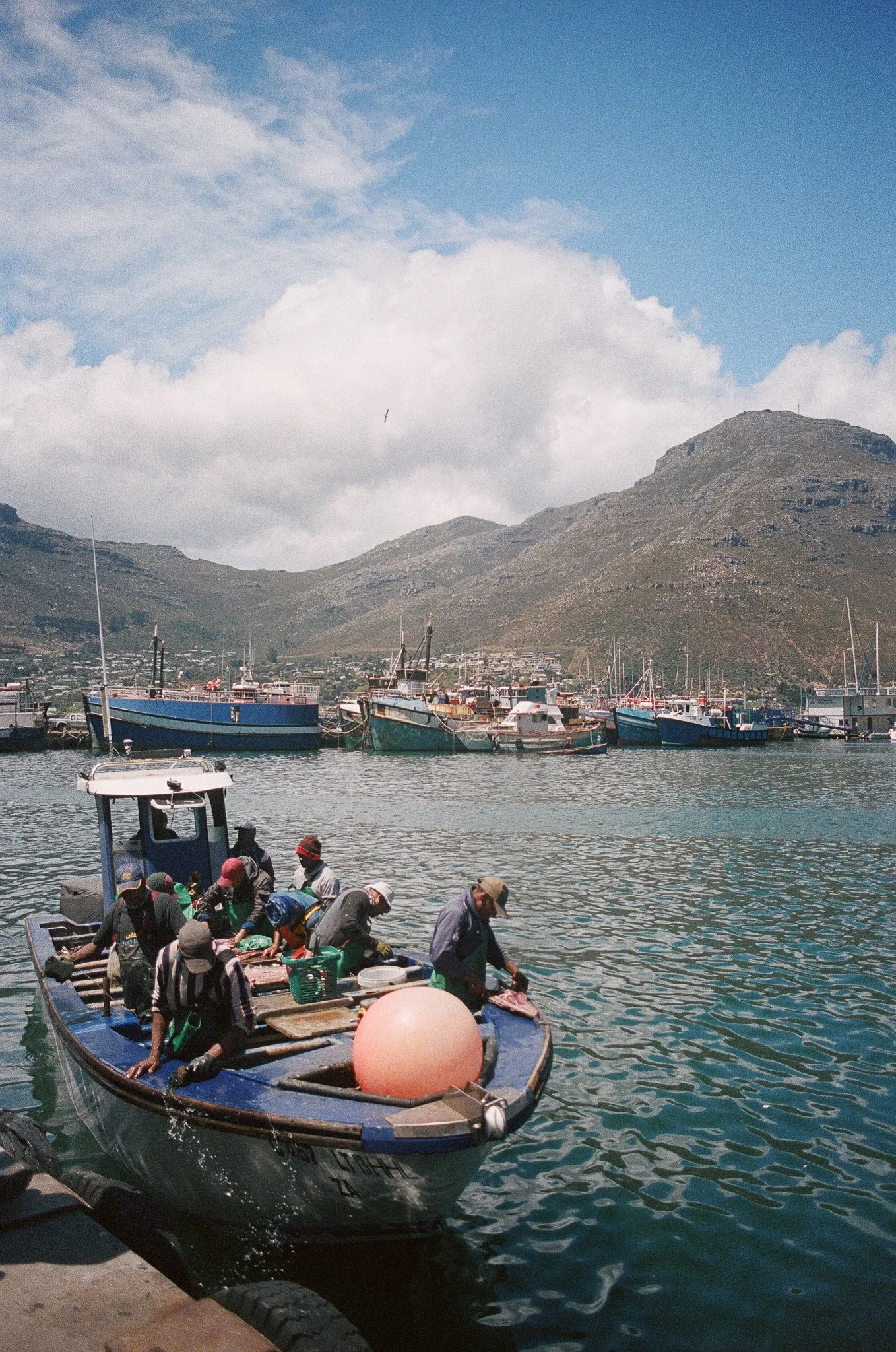 A group of people on a boat docked at a marina, with mountains and boats in the background, under a partly cloudy sky.