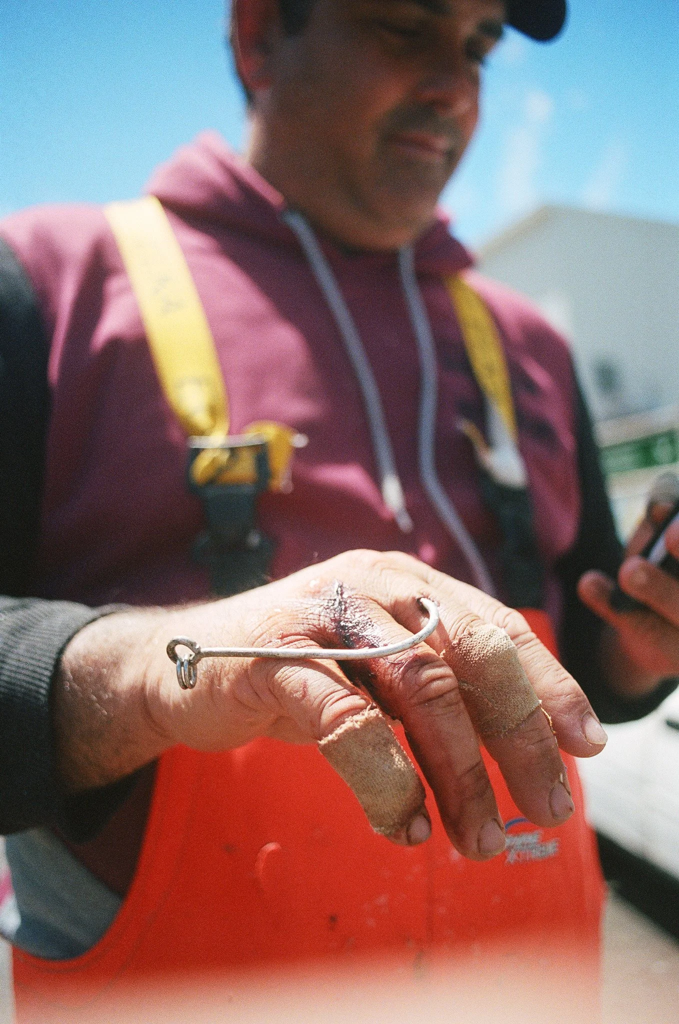 Close-up of a person's hand holding a fish hook with fish flesh, with a person wearing a pink hoodie and yellow strap in the background.