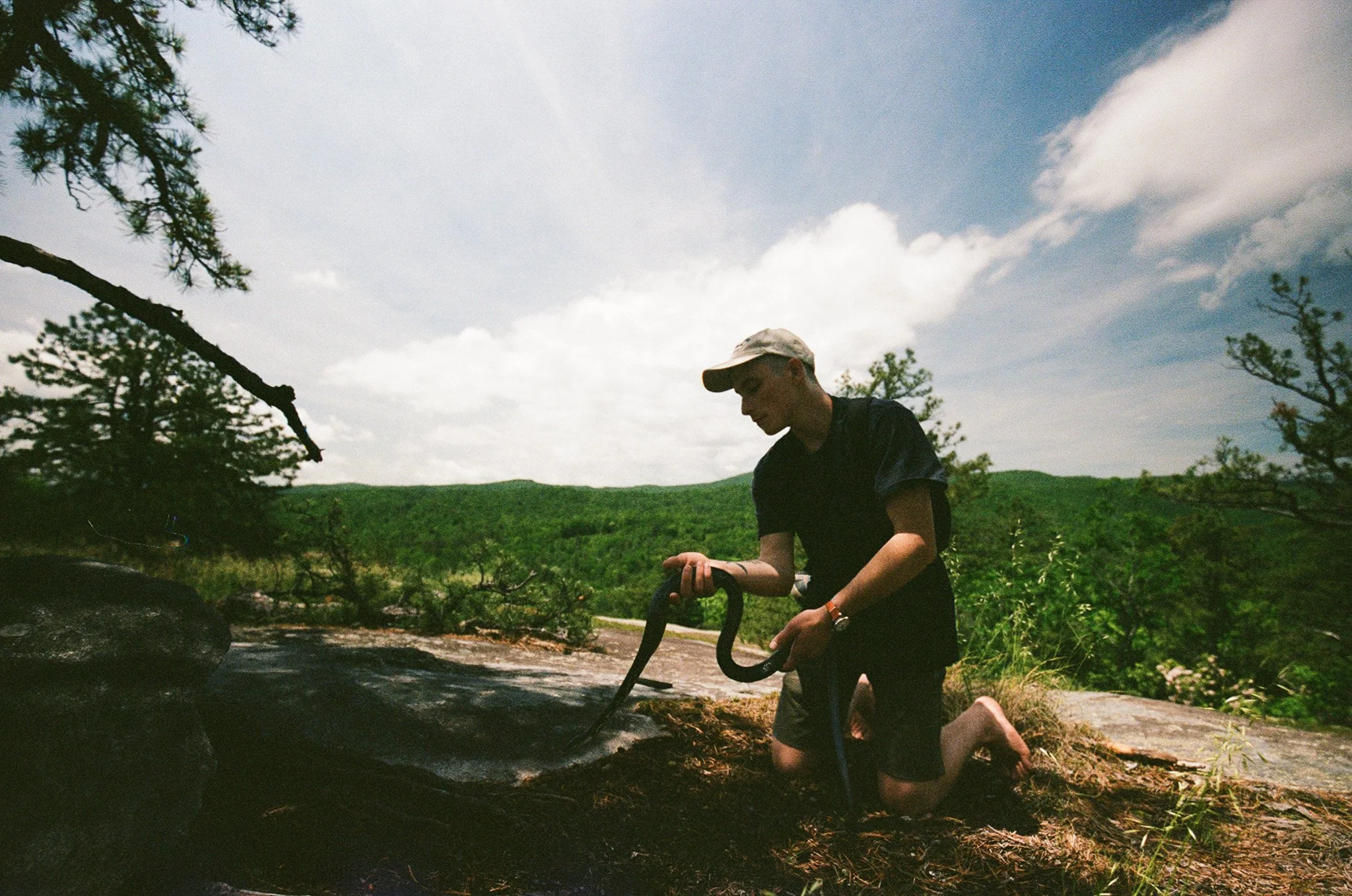 A person kneeling on the ground outdoors, using a tool to work on a small fire or campfire, with trees and a cloudy sky in the background.
