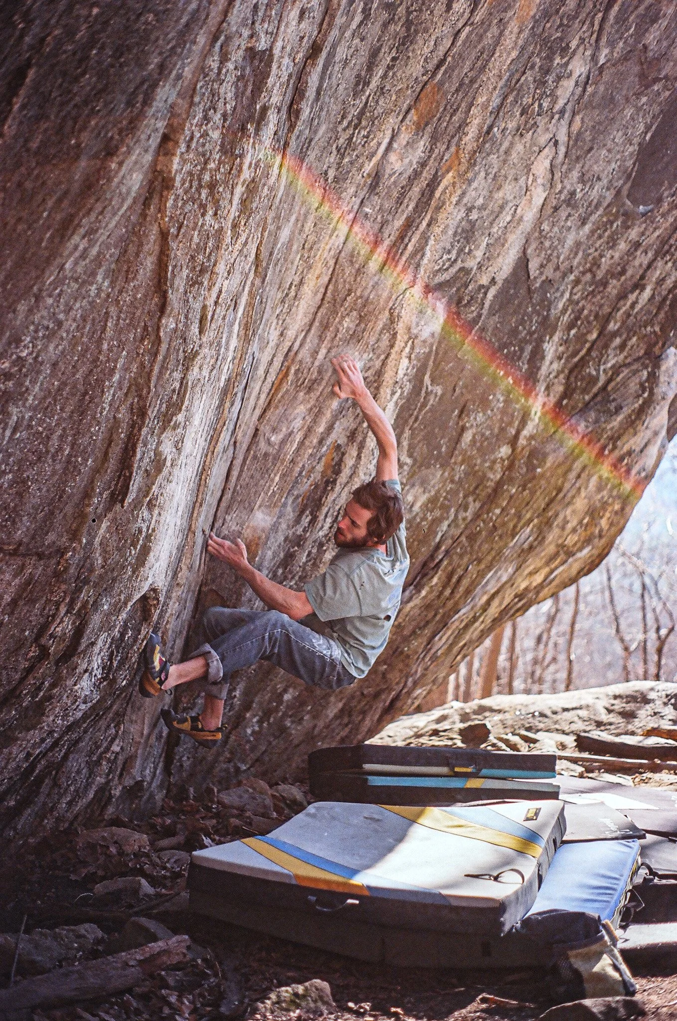 A person indoor rock climbing on a vertical wooden climbing wall with a rainbow lens flare across the image, and climbing mats on the ground below.
