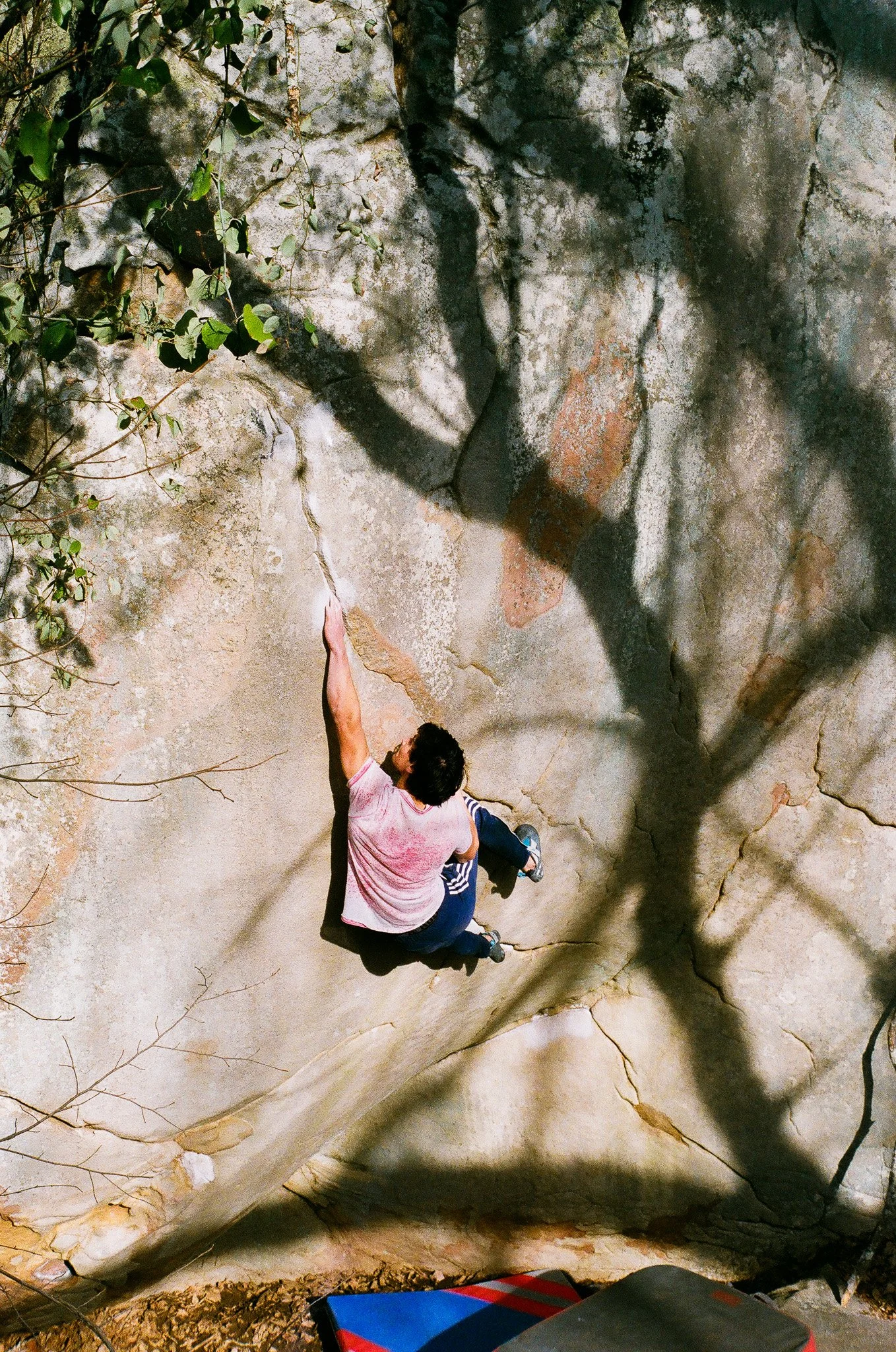 A person bouldering on a large, weathered rock surface outdoors, reaching upward with one hand, wearing a pink shirt, dark pants, and climbing shoes, surrounded by natural scenery and shadows.