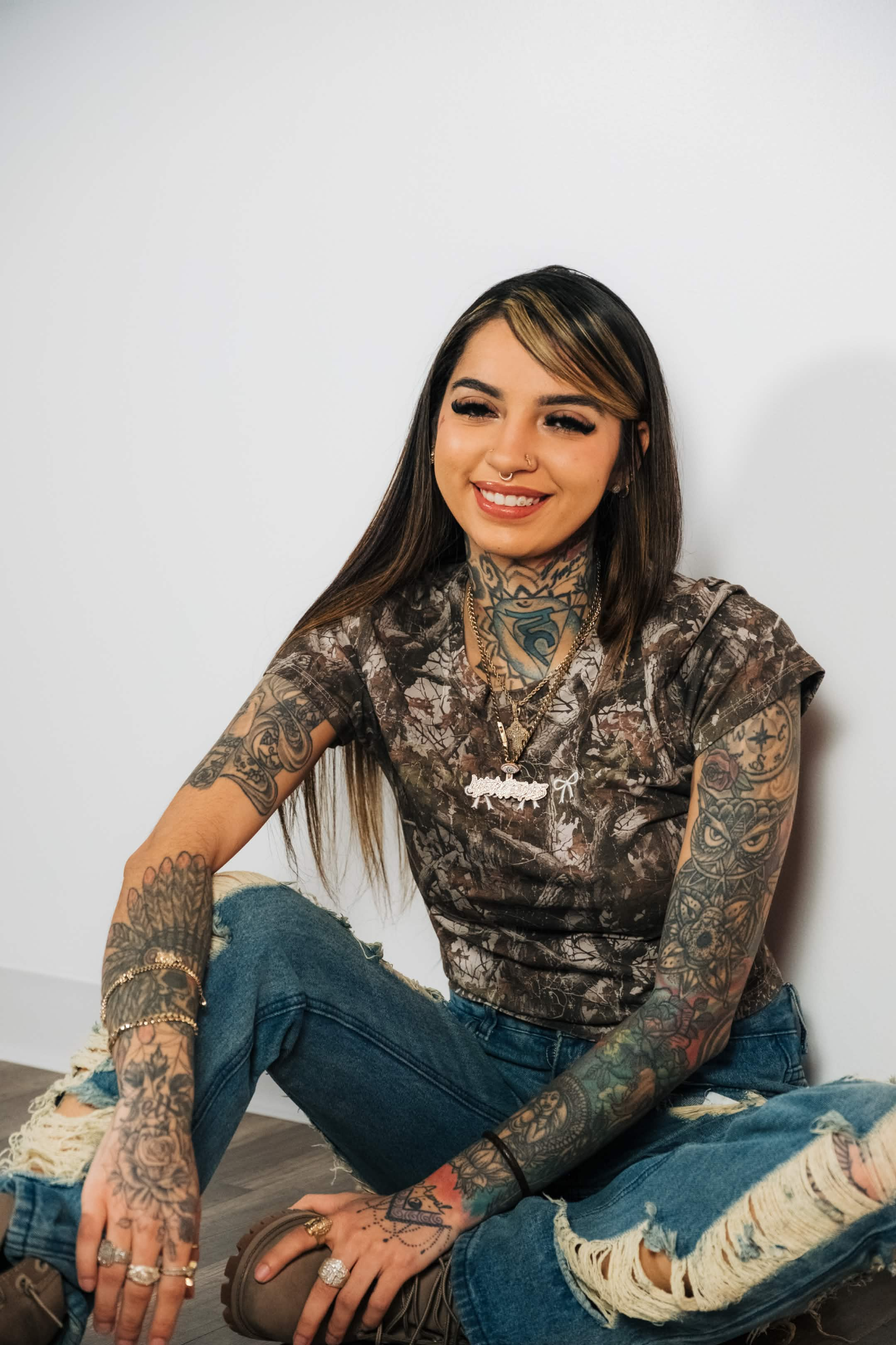 A woman with long dark hair and tattoos on her arms and neck sitting on the floor in front of a plain white wall, smiling with closed eyes, dressed in a camouflage-patterned shirt, ripped jeans, and wearing jewelry.