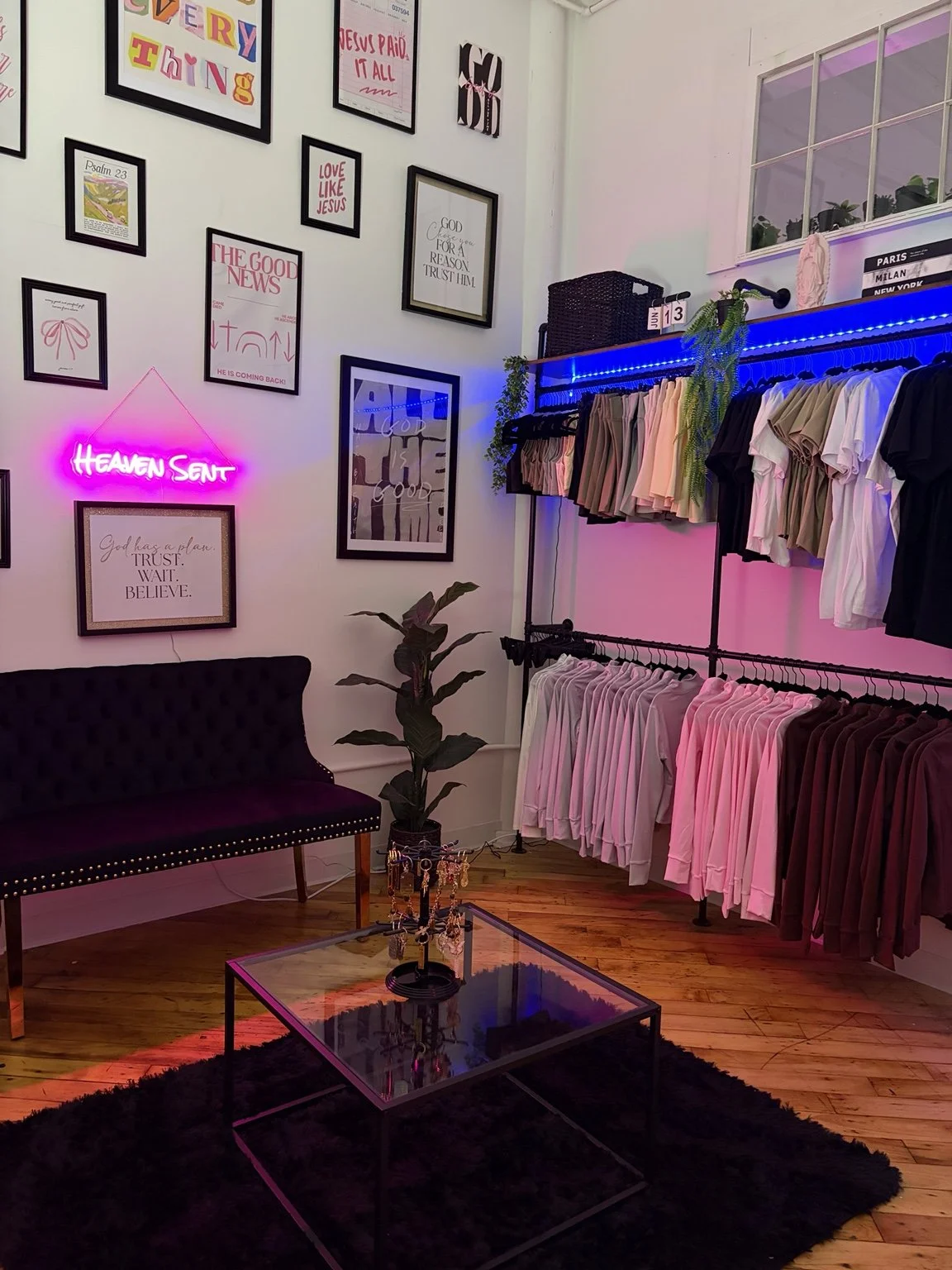 A small boutique clothing store with a black velvet sofa, a glass coffee table, and a wall decorated with framed inspirational quotes and artwork. There is a neon sign that says 'HEAVEN SENT' in pink, and clothing racks holding pastel and neutral-colored shirts and pants. The store has pink and blue LED lighting accents and a potted plant near the wall.