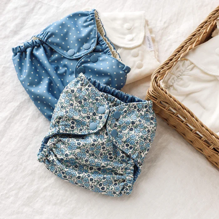 Two cloth diapers with snaps, one blue with white polka dots and the other with a blue floral pattern, placed on a light-colored surface, next to a wicker basket containing folded cloth diapers.