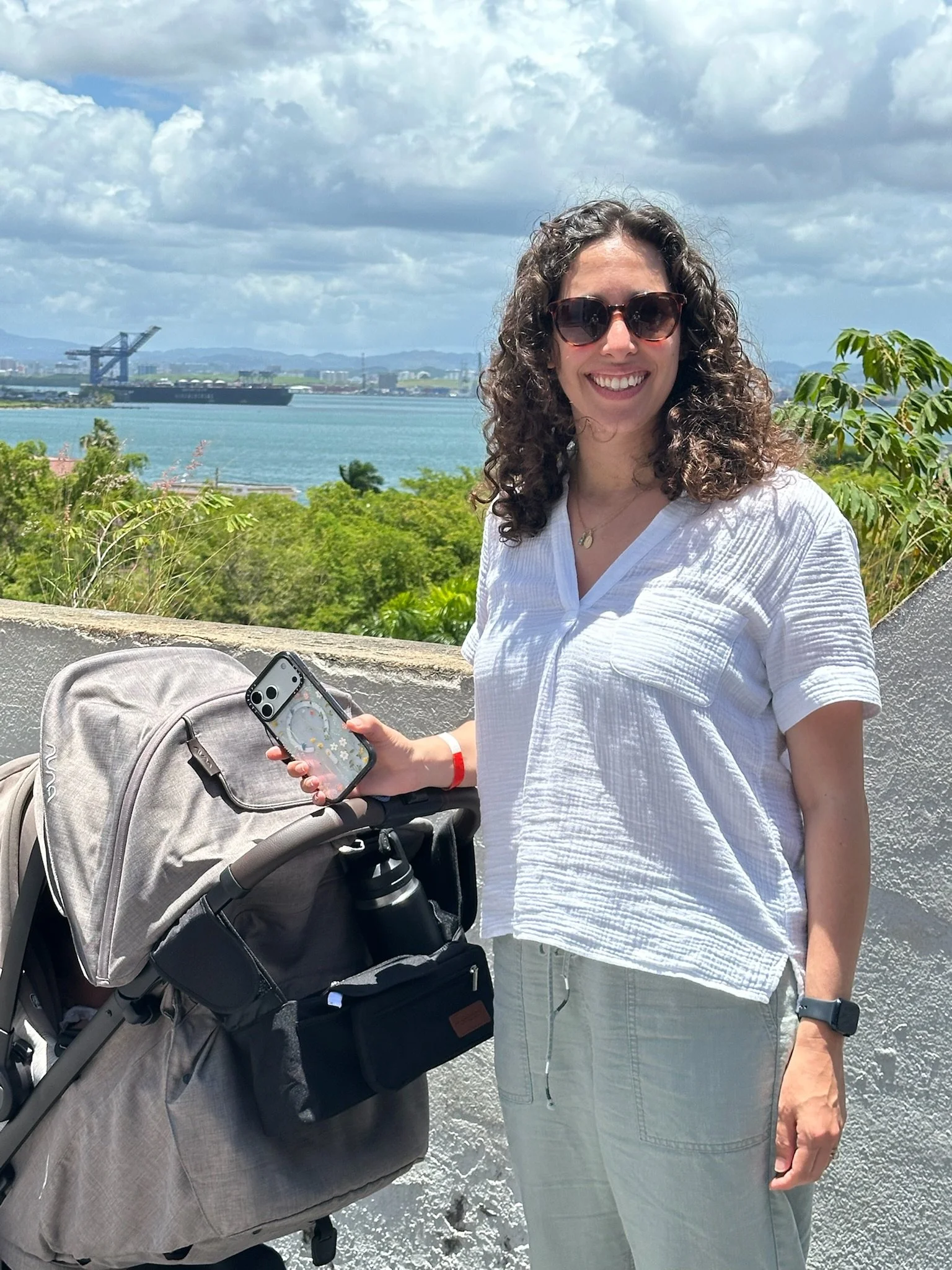 A woman with curly hair and sunglasses smiling outdoors, holding a smartphone, standing next to a stroller with a bag, with a body of water, a ship, and a cloudy sky in the background.