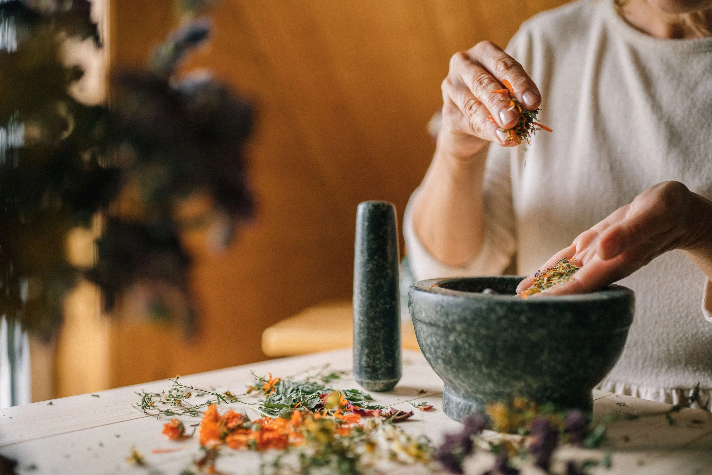 A person with long hair, wearing a beige shirt, is using their hands to prepare herbs or flowers over a black bowl, with herbs and flowers scattered on a white table.