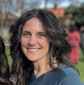 Portrait of a woman with curly dark hair, smiling outdoors in a park with blurred background of trees and people.