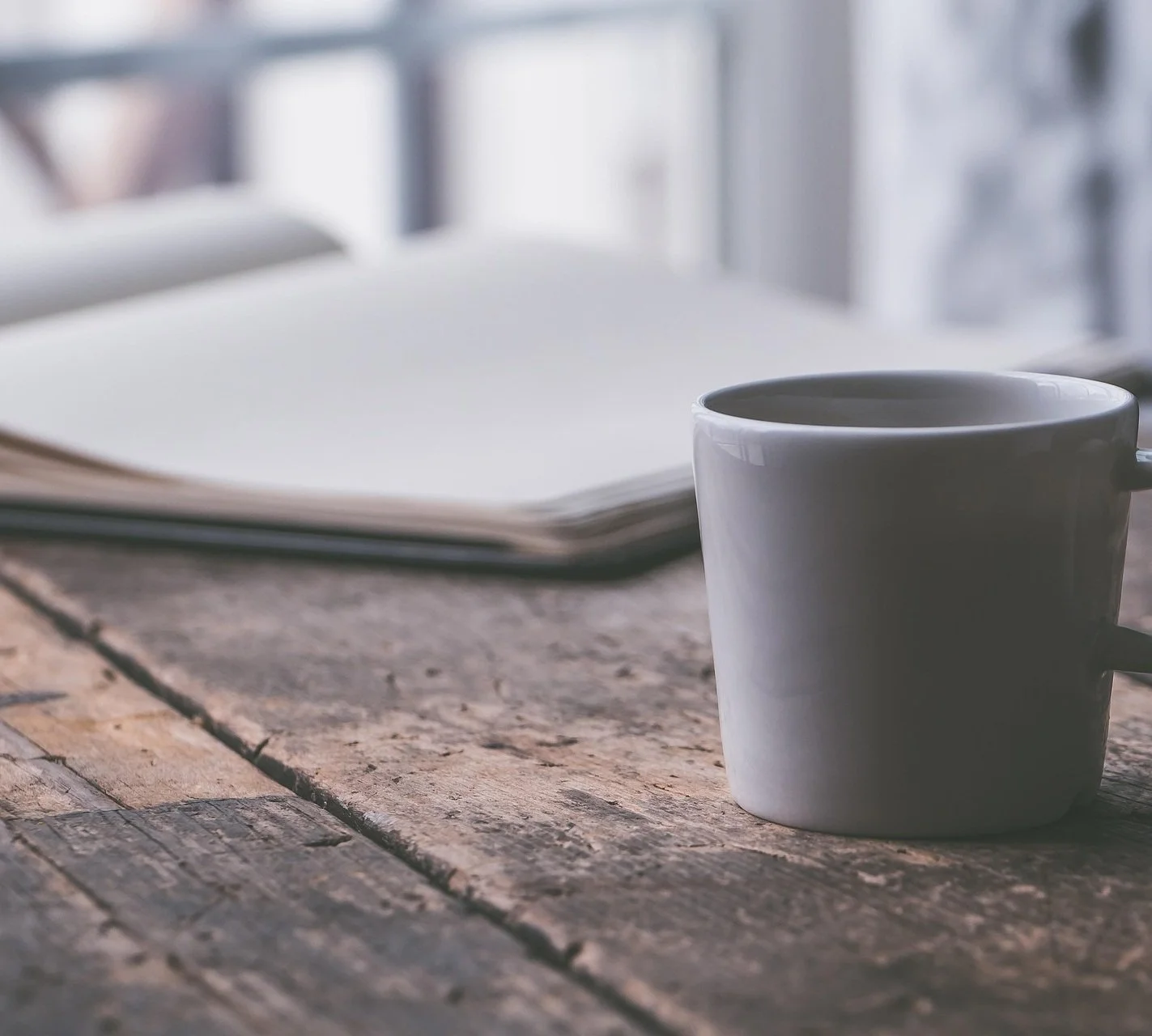 Close-up of a white coffee mug on a rustic wooden table, with an open notebook or journal in the background, near a window.