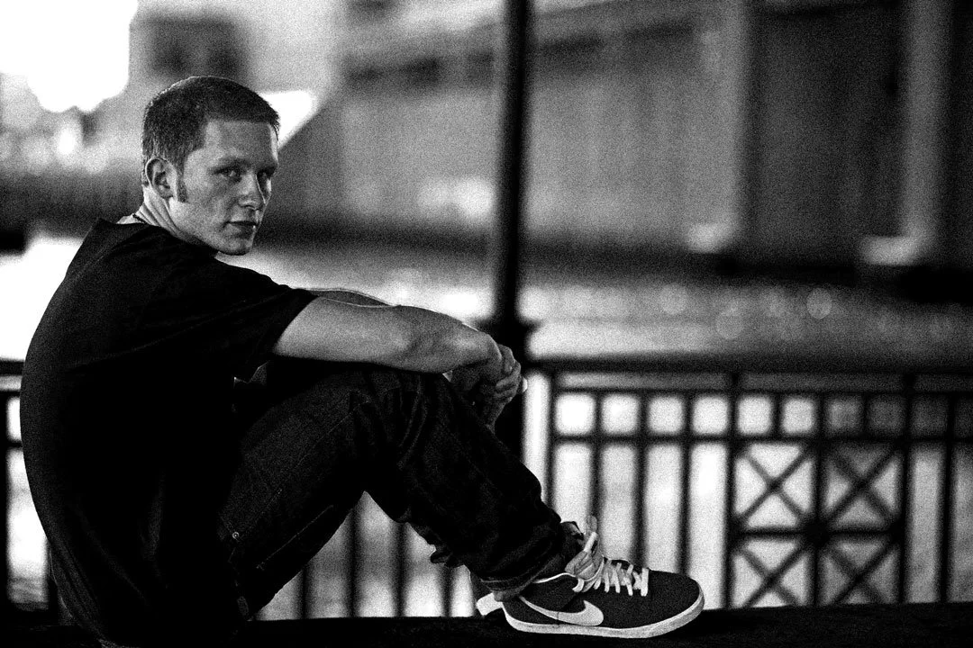 A young man sitting on a ledge at night, wearing a dark t-shirt, jeans, and Nike sneakers, with a railing and blurred city lights in the background.