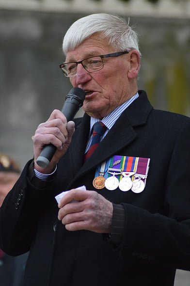 An elderly man with gray hair and glasses, wearing a dark suit jacket with military medals, holding a microphone and reading from a piece of paper during a ceremony.