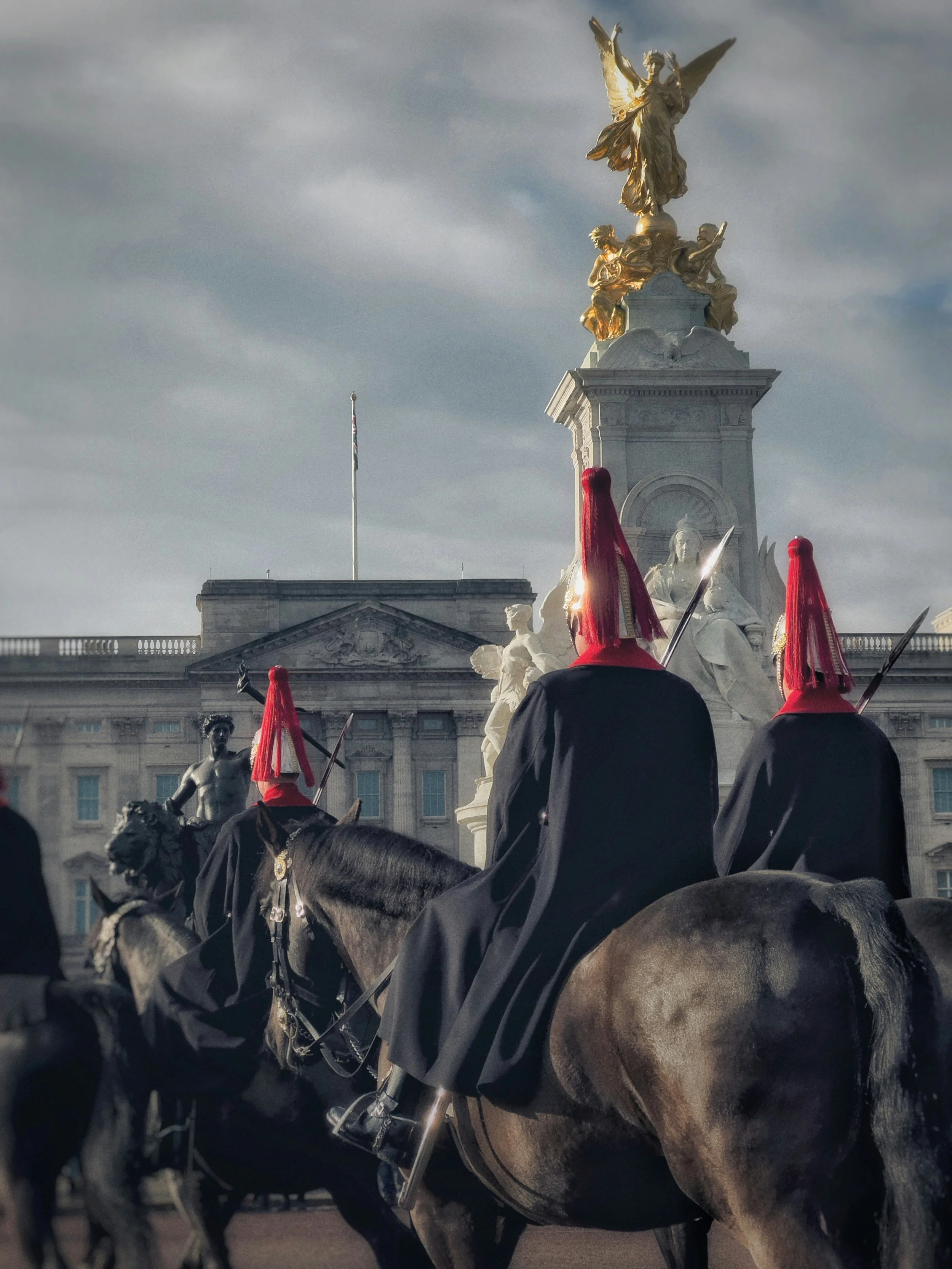 A group of mounted soldiers dressed in black with red headgear and spears, in front of a large historic building with an ornate statue above.