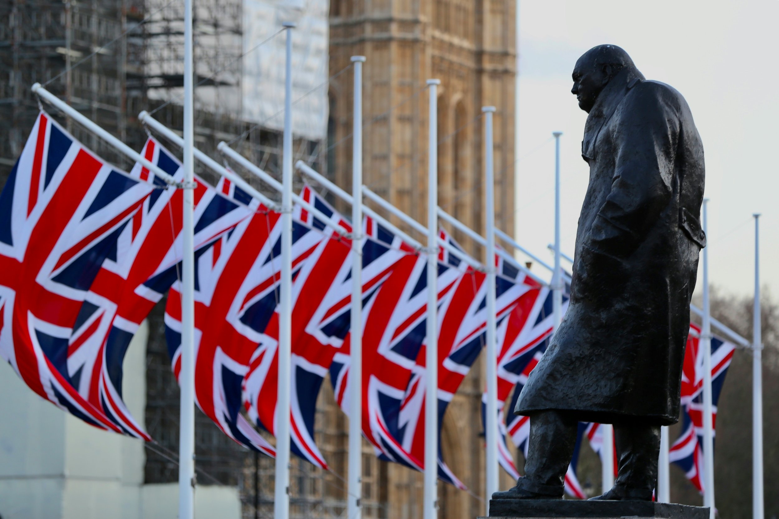 A statue of a man in a suit near Union Jack flags, with a large historic building under construction or renovation in the background.