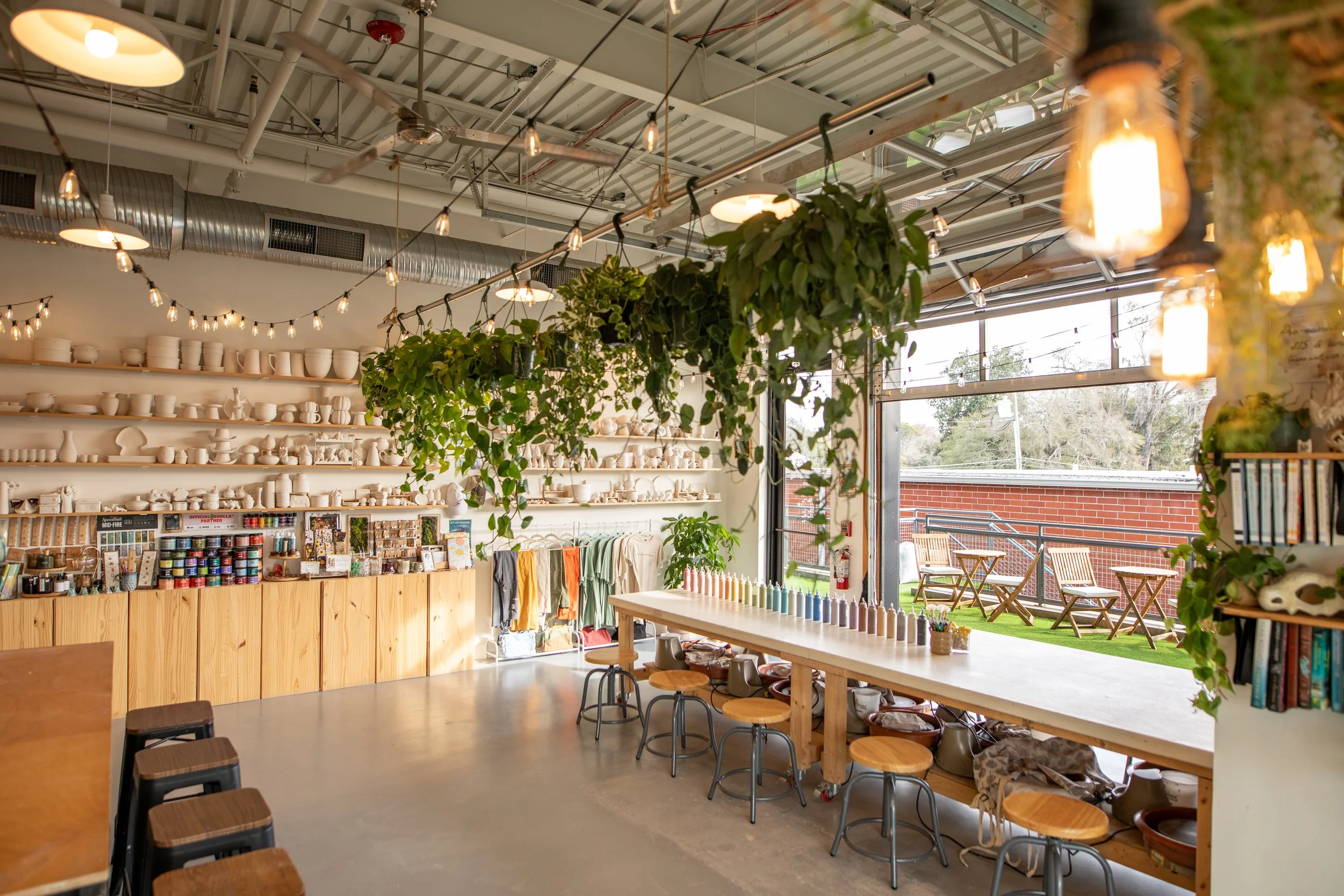 Interior of a retail store with wooden shelves filled with pottery, shelves with yarn, and large windows letting in natural light, decorated with hanging string lights and leafy plants.