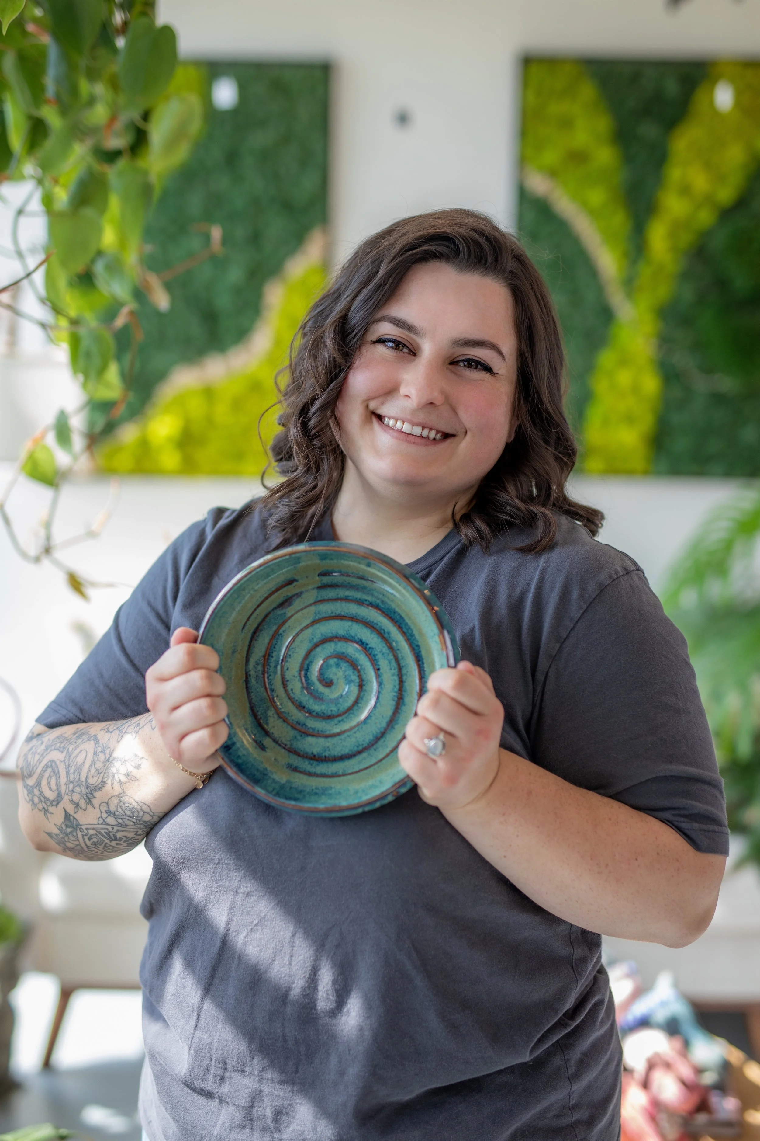 A woman with dark hair, smiling, holding up a ceramic plate with a blue and green spiral design. There are plants and artwork in the background.